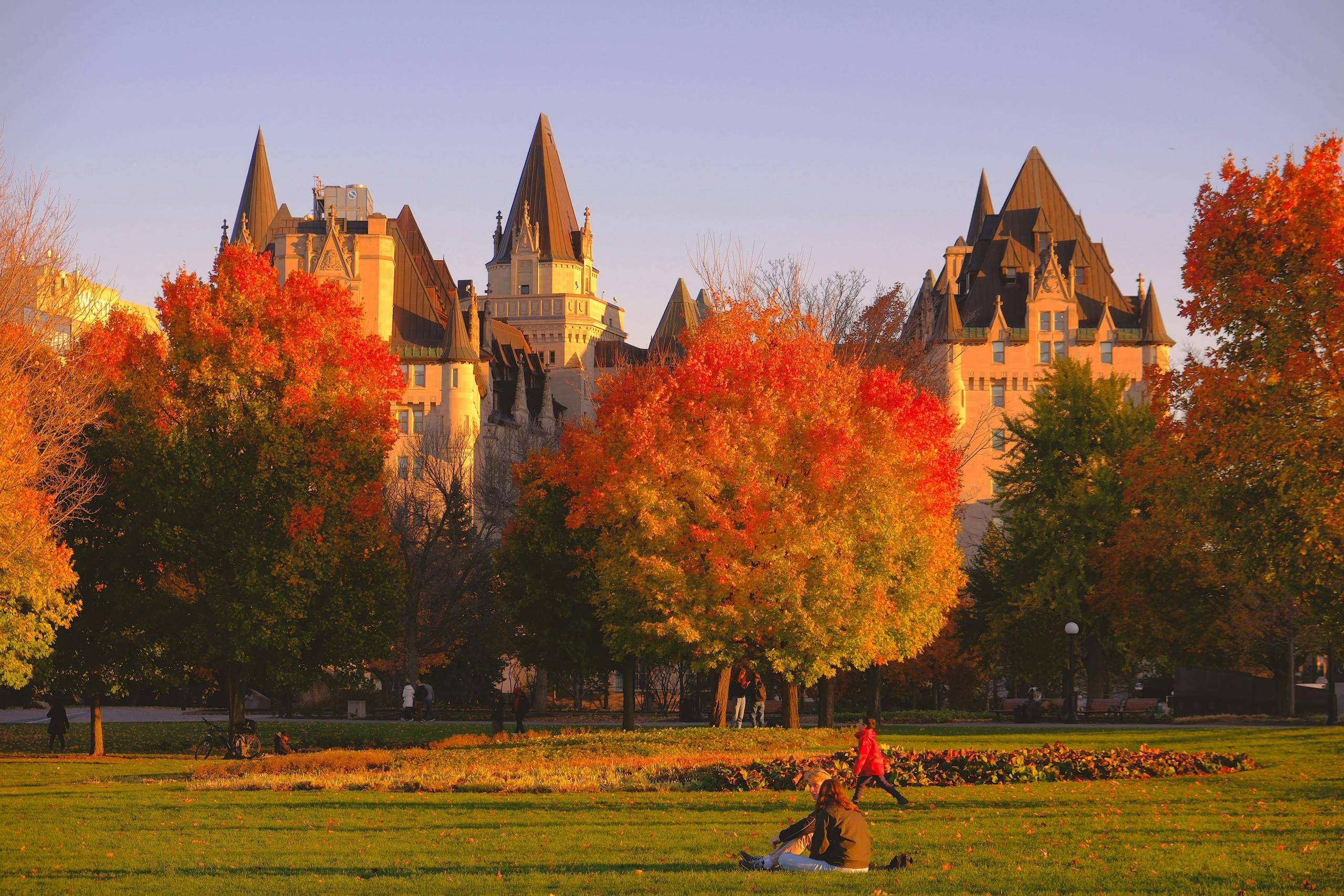 People enjoying a sunny autumn day in a park in Ottawa, surrounded by fall foliage with the Château Laurier in the background.