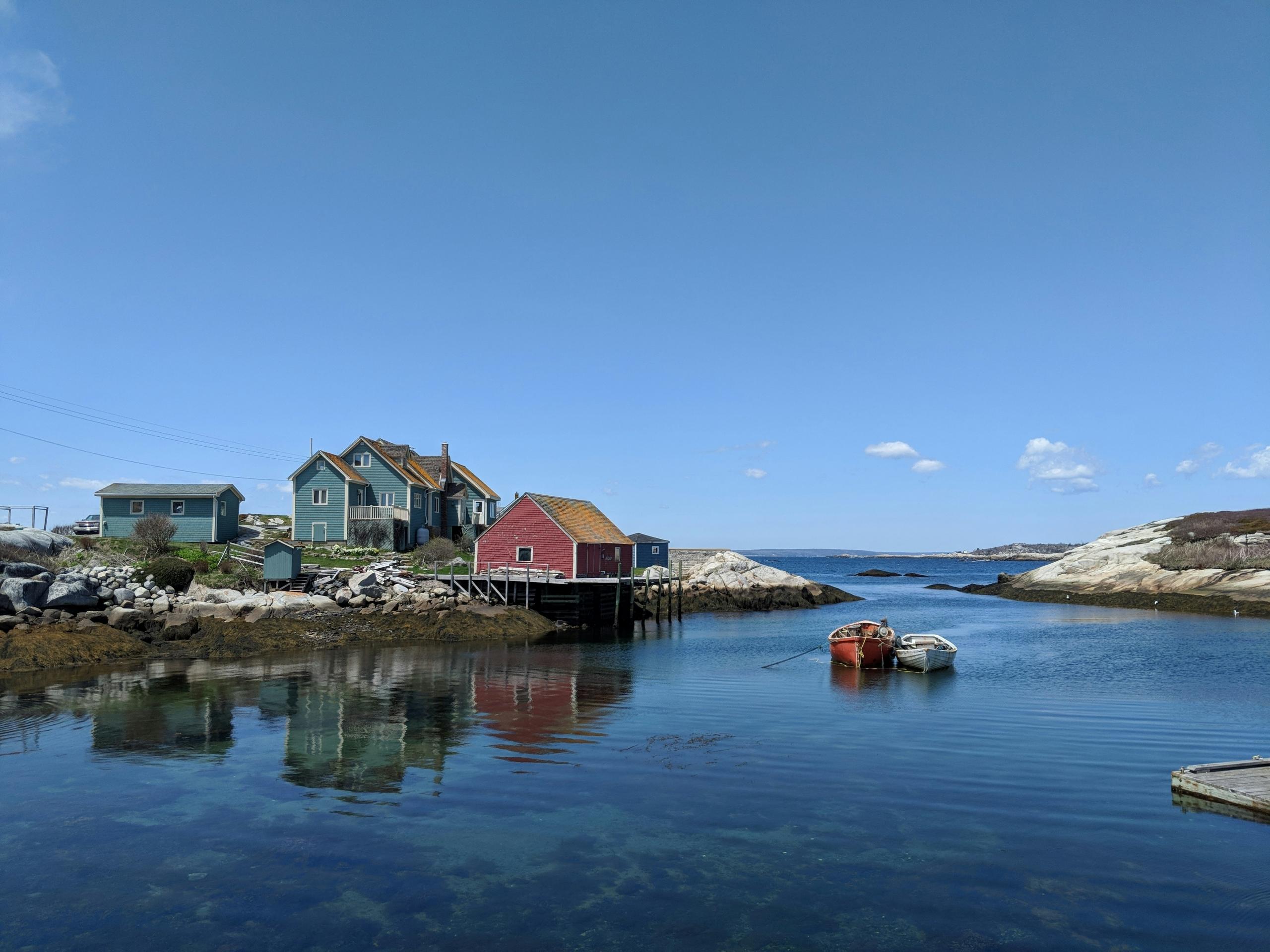 A red boathouse and green homes reflect in the clear, calm water of the harbor. Two small boats float nearby, and rocky outcrops frame the serene blue sea under a bright, clear sky.