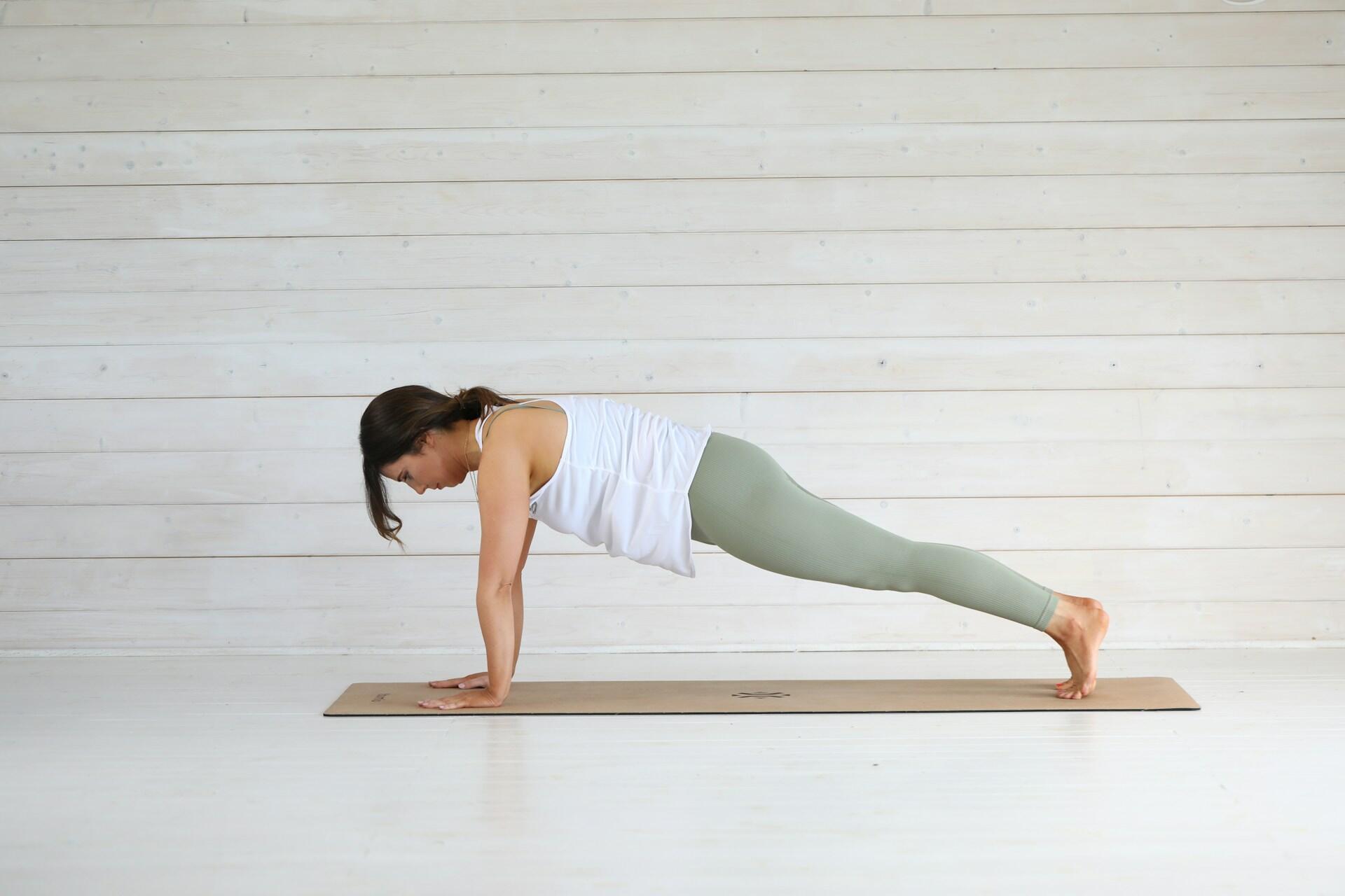 Woman in white tank top and green leggings holding a plank pose on a yoga mat against a light wooden wall background.