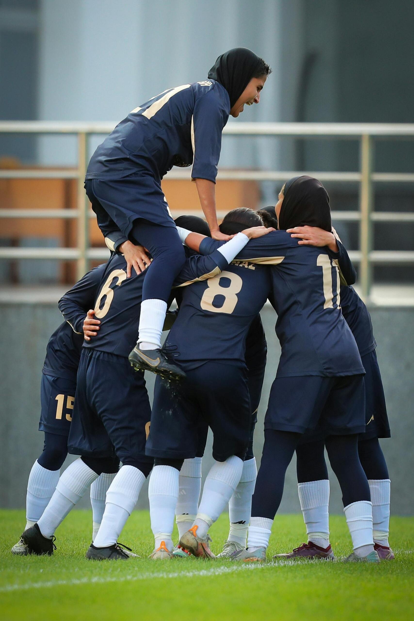 A group of female soccer players wearing navy blue uniforms and hijabs celebrating on the field with one player lifted above the others.