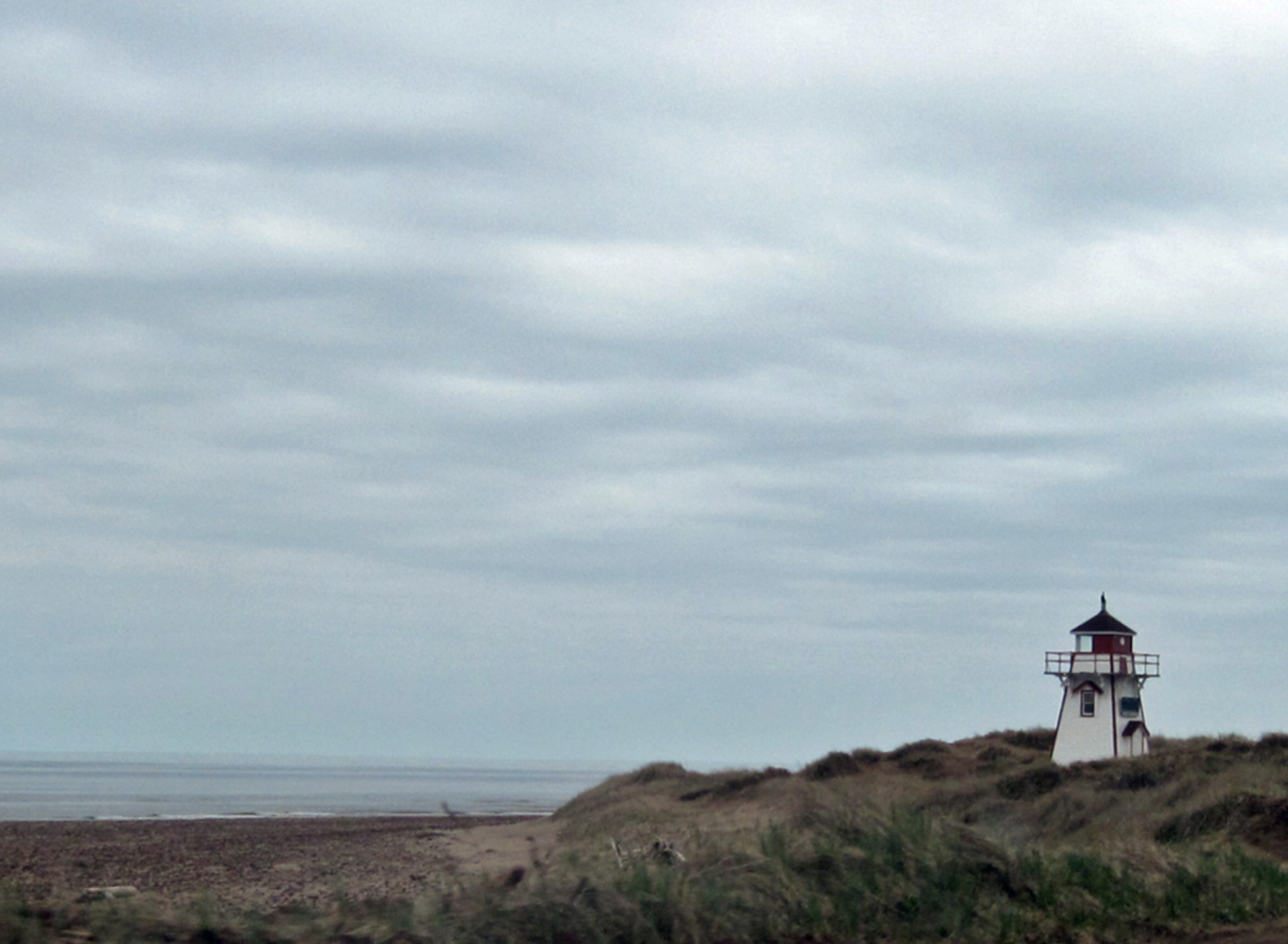 A small lighthouse stands alone on a grassy dune overlooking a vast, rocky beach and calm sea under a cloudy sky. The lighthouse, painted white with red accents, features a black roof and a wraparound balcony. The surrounding landscape is sparse and rugged, with the muted colors of the overcast day creating a serene and slightly melancholic atmosphere.