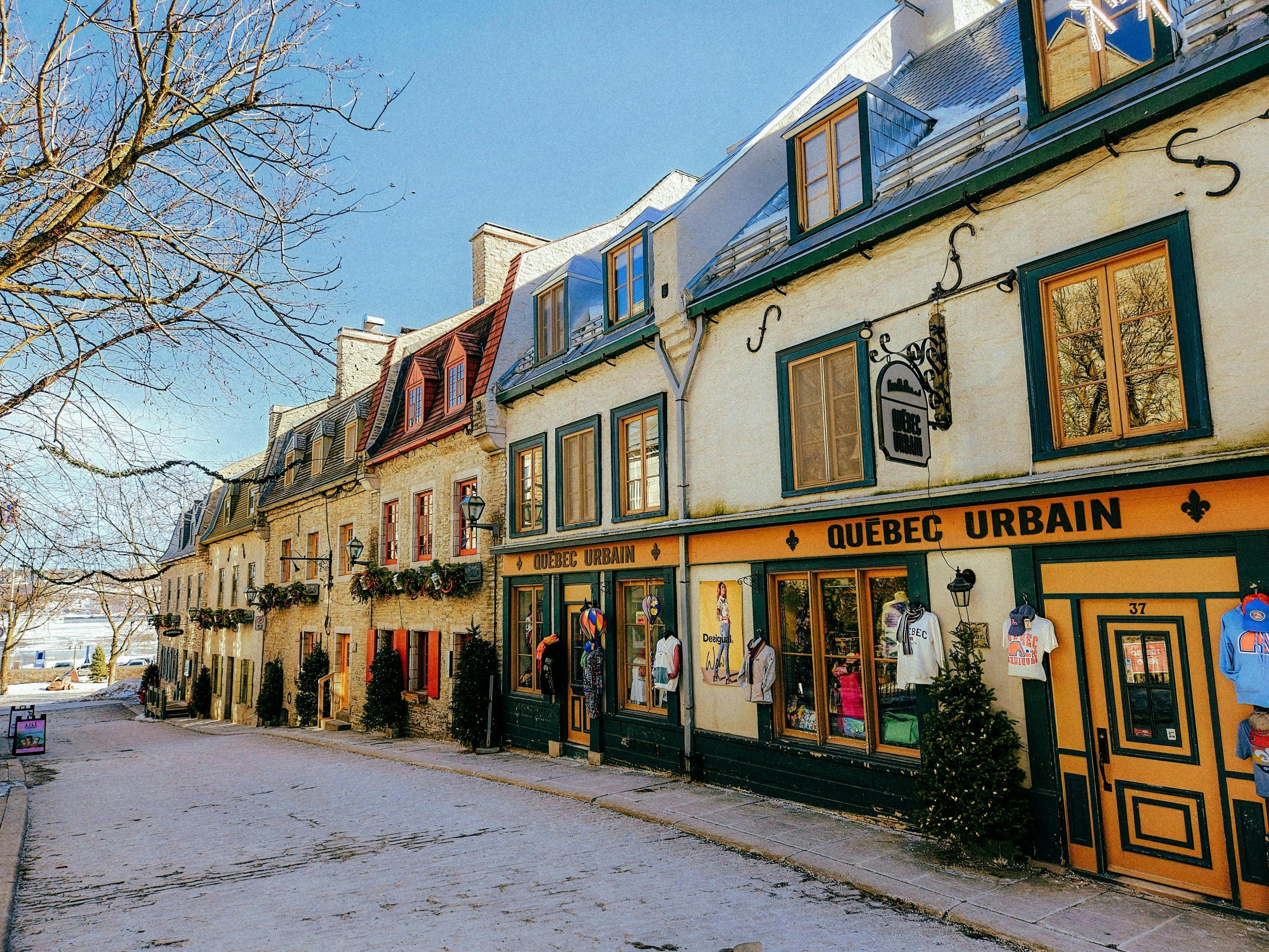 A street in Quebec City's Old Town, featuring historic stone buildings with colorful windows and storefronts. The shop "Quebec Urbain" displays clothing items outside. Leafless trees line the cobblestone street under a clear blue sky, capturing a quaint and picturesque winter scene.