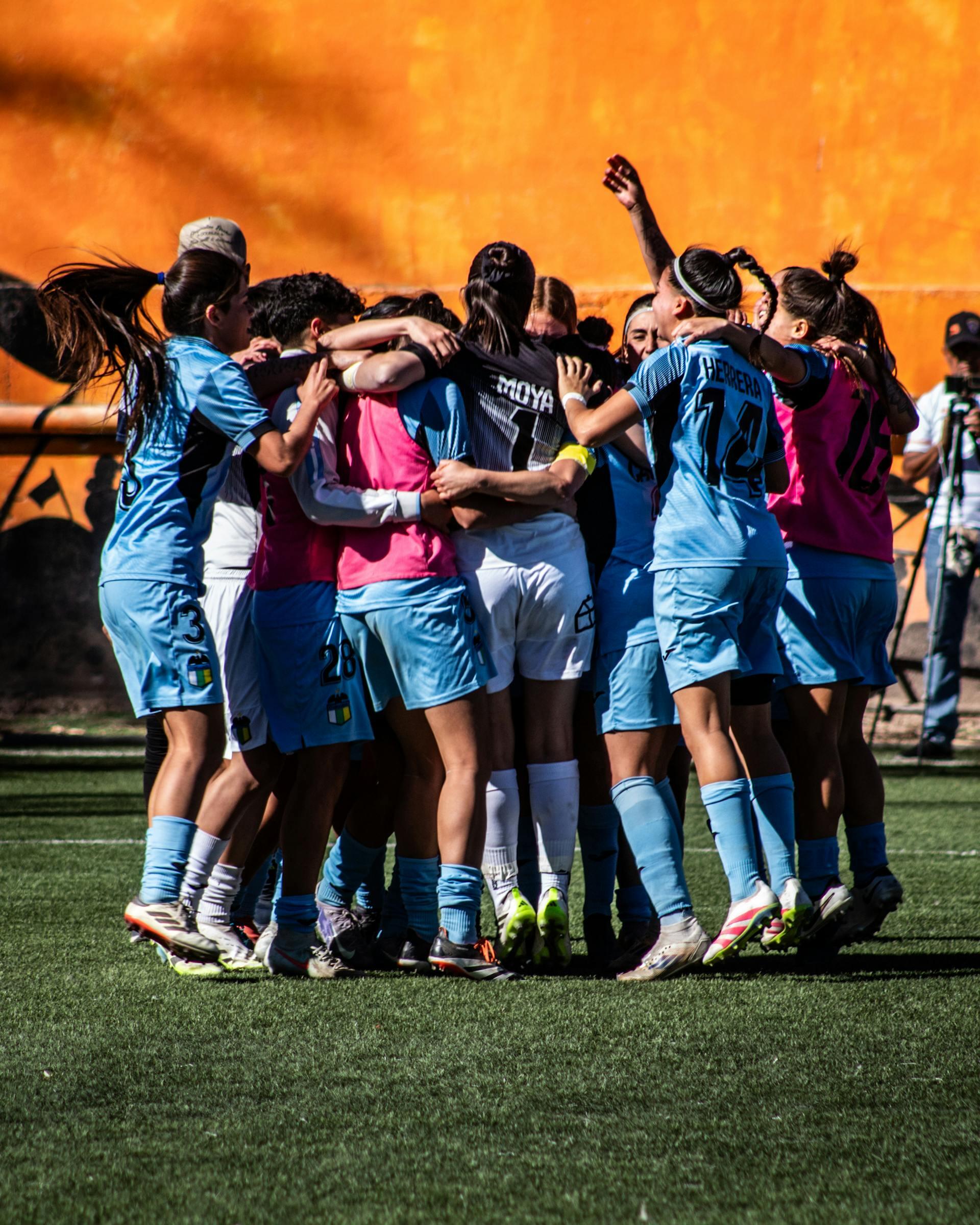 Women soccer players in light blue uniforms huddled together celebrating on a green field with an orange wall background.