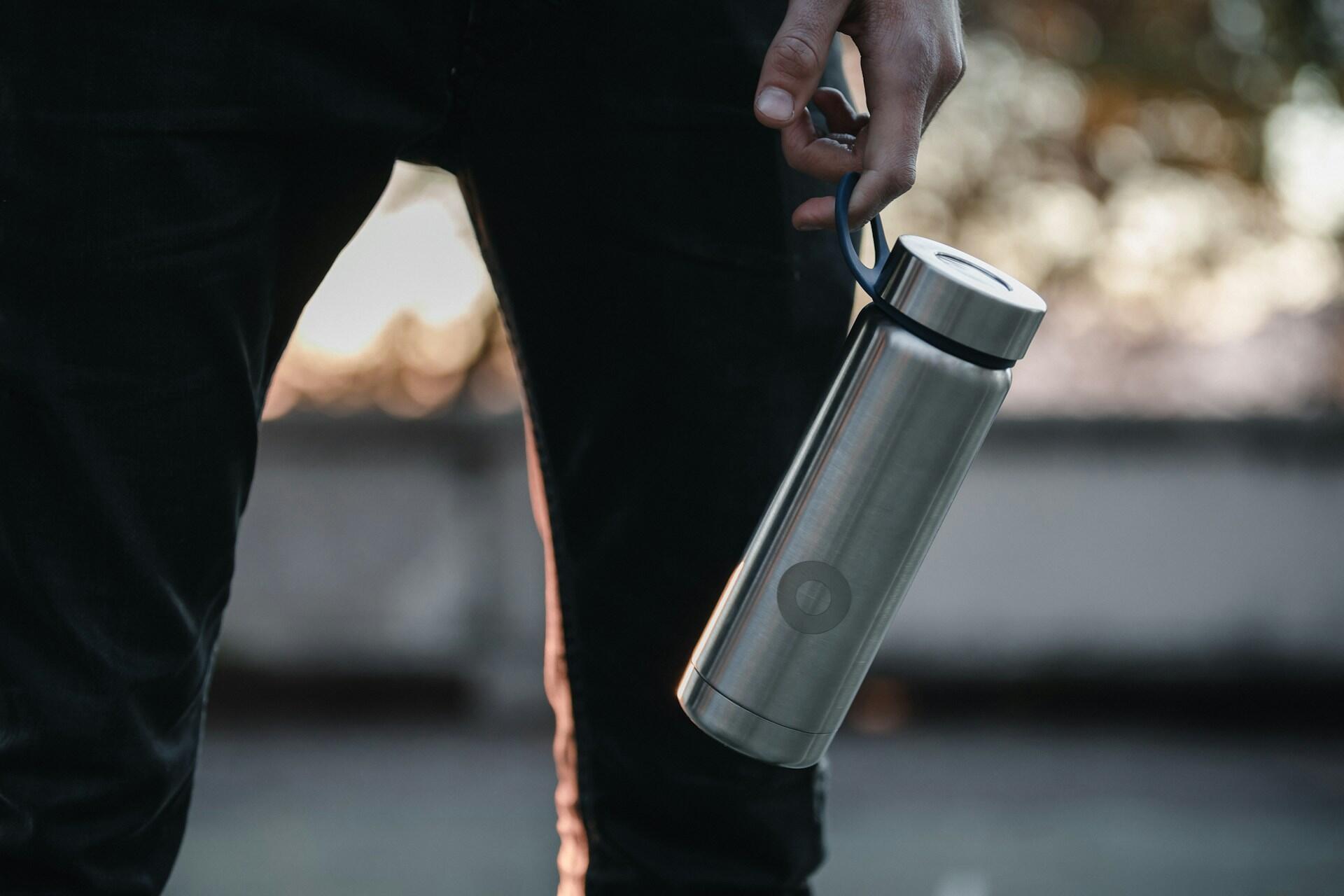Person holding a stainless steel insulated water bottle by its handle loop, standing outdoors with blurred background.