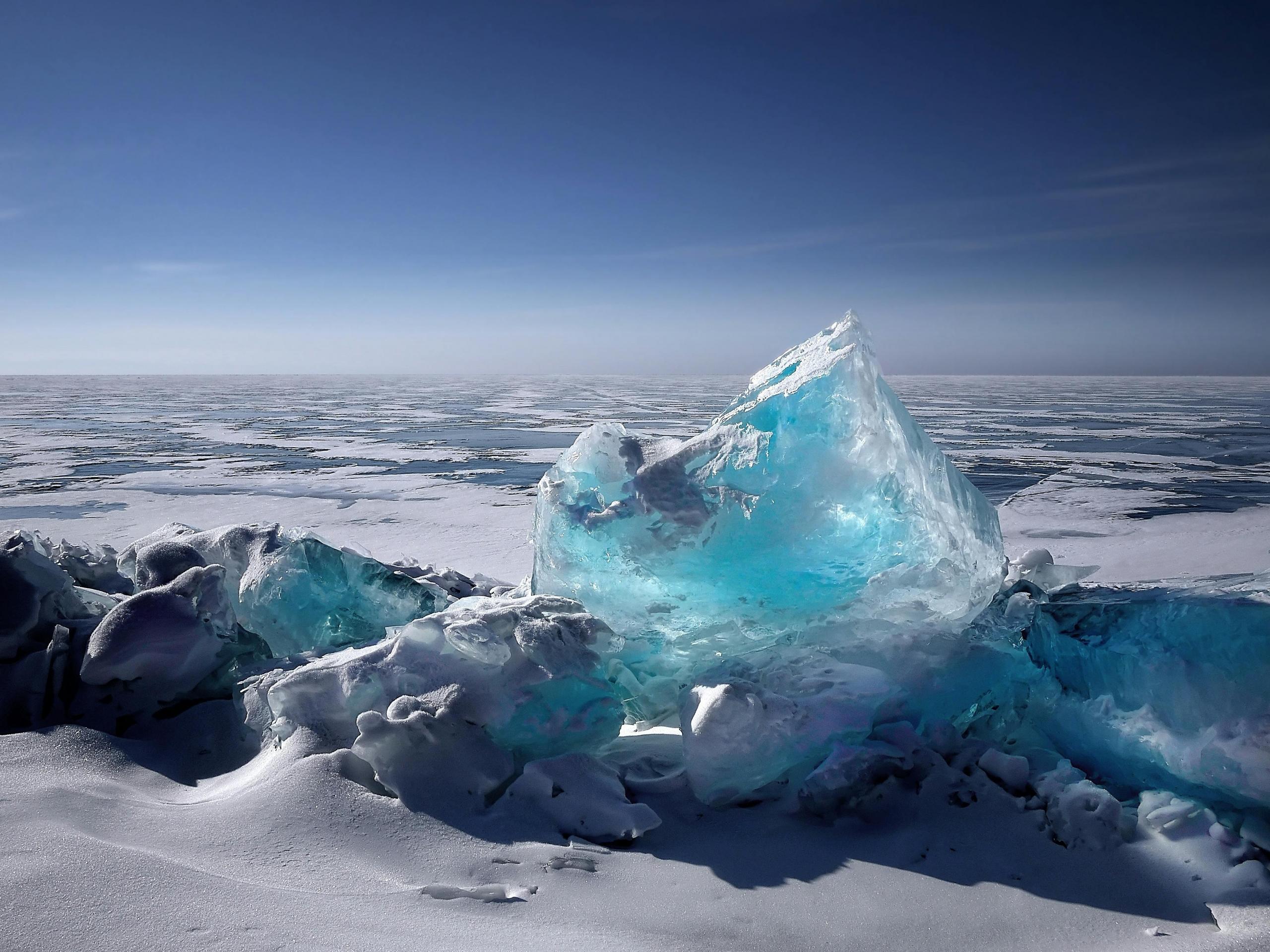 A stunning Arctic landscape featuring a large, jagged piece of blue ice standing prominently against a vast expanse of snow and ice. The clear blue sky above contrasts sharply with the icy terrain.