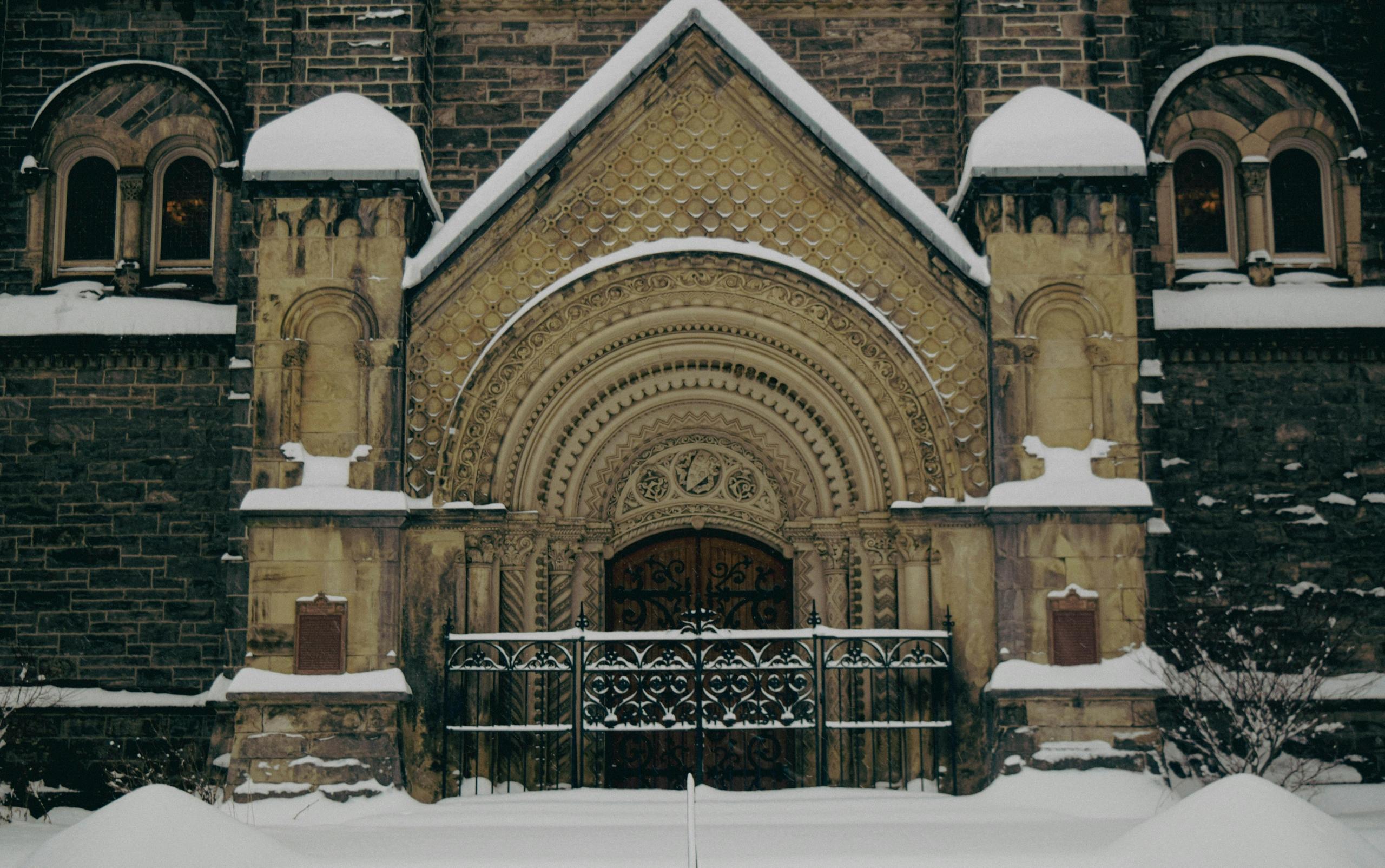 The grand entry to the university of Toronto. Snow covers the elaborate doorway to the university. Snow covers the ground. A gothic fence is in front of wooden doors.