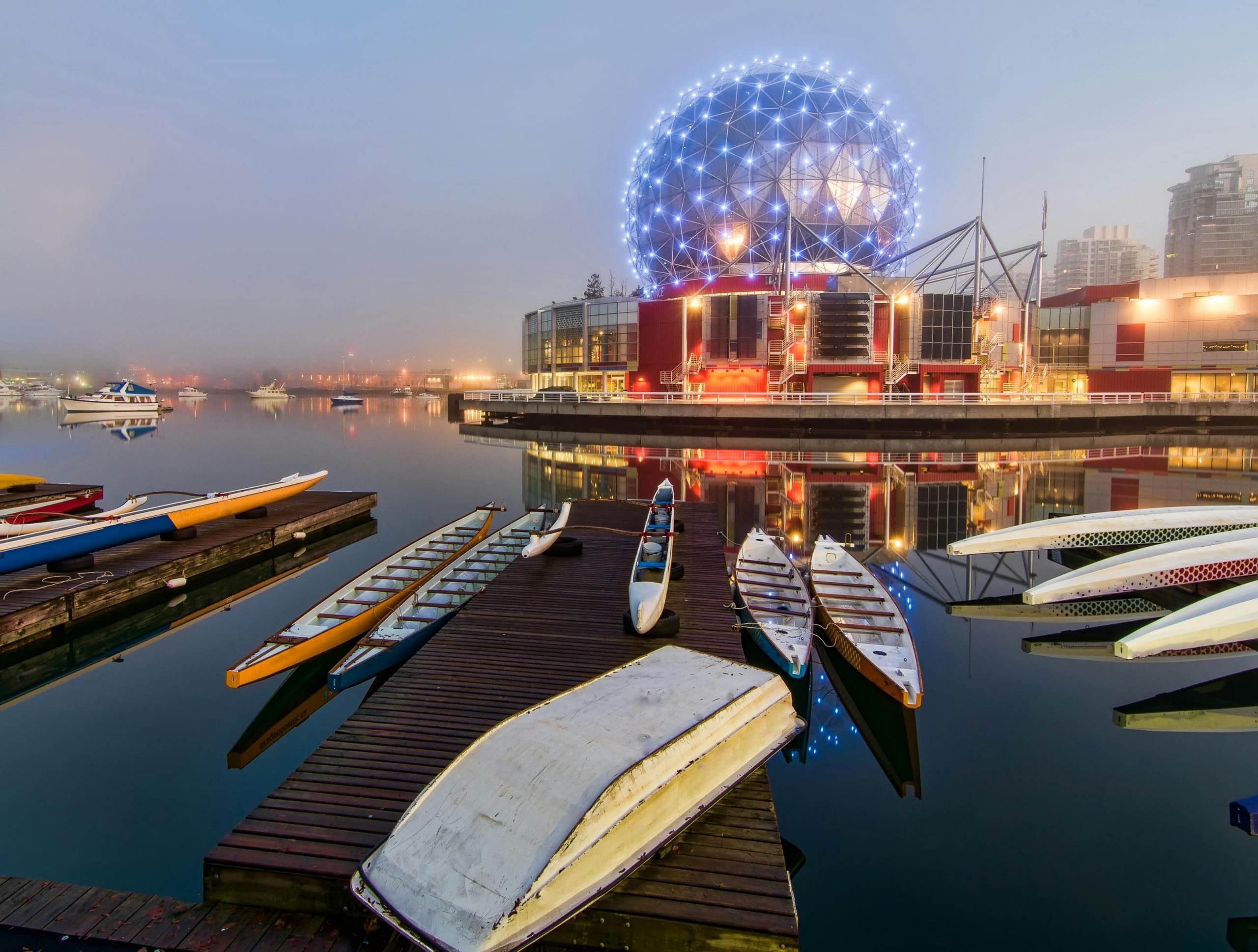 An evening view of False Creek in Vancouver, British Columbia, showcasing brightly colored kayaks and canoes docked at a wooden pier. In the background, the illuminated geodesic dome of Science World reflects on the calm water, with the city skyline faintly visible through the mist.