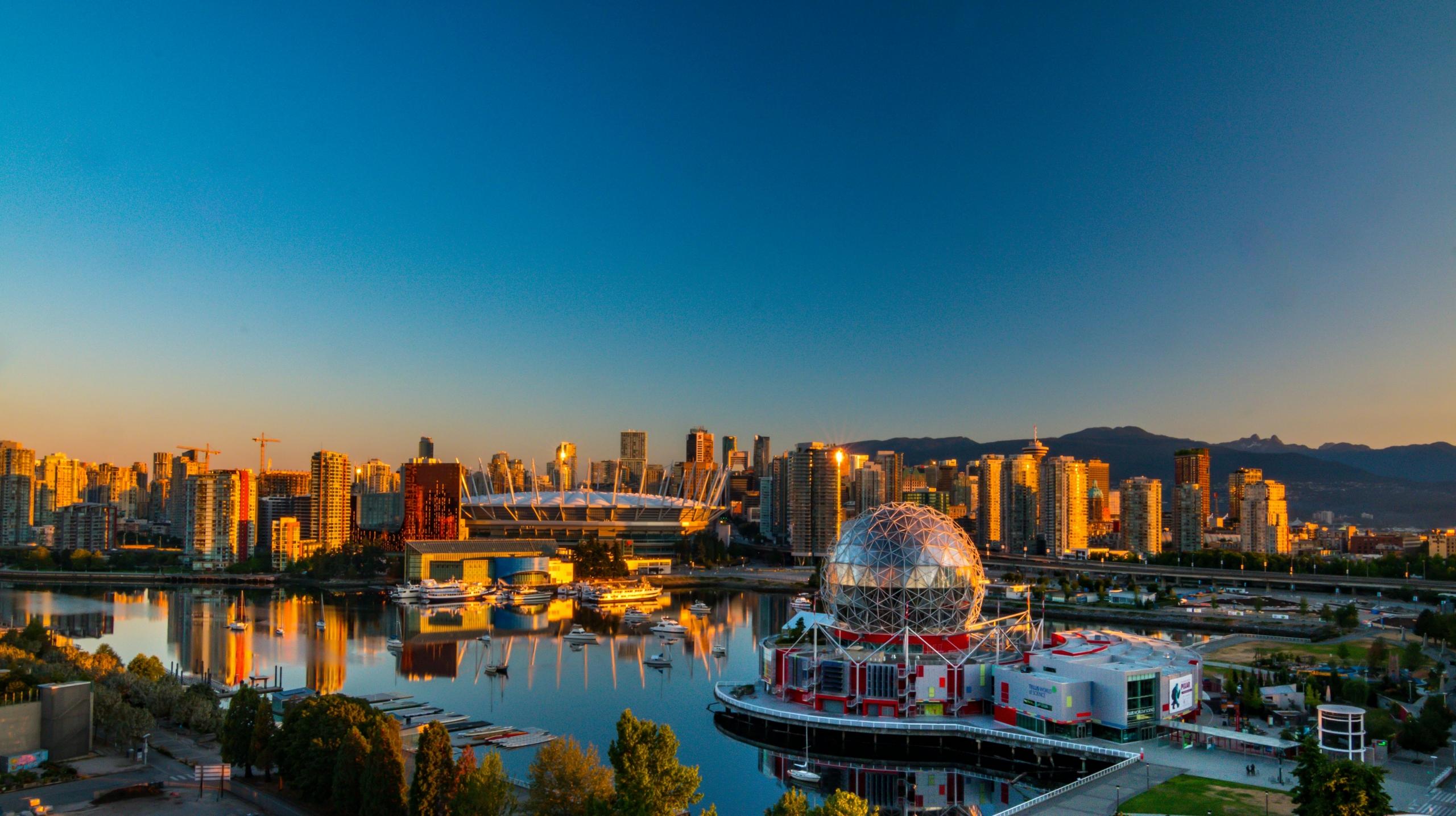 A cityscape of Vancouver, British Columbia, at sunset. The skyline features high-rise buildings reflecting on the water, with mountains in the background. The BC Place Stadium and the geodesic dome of Science World are visible. The sky is clear, with a gradient from blue to warm hues as the sun sets.