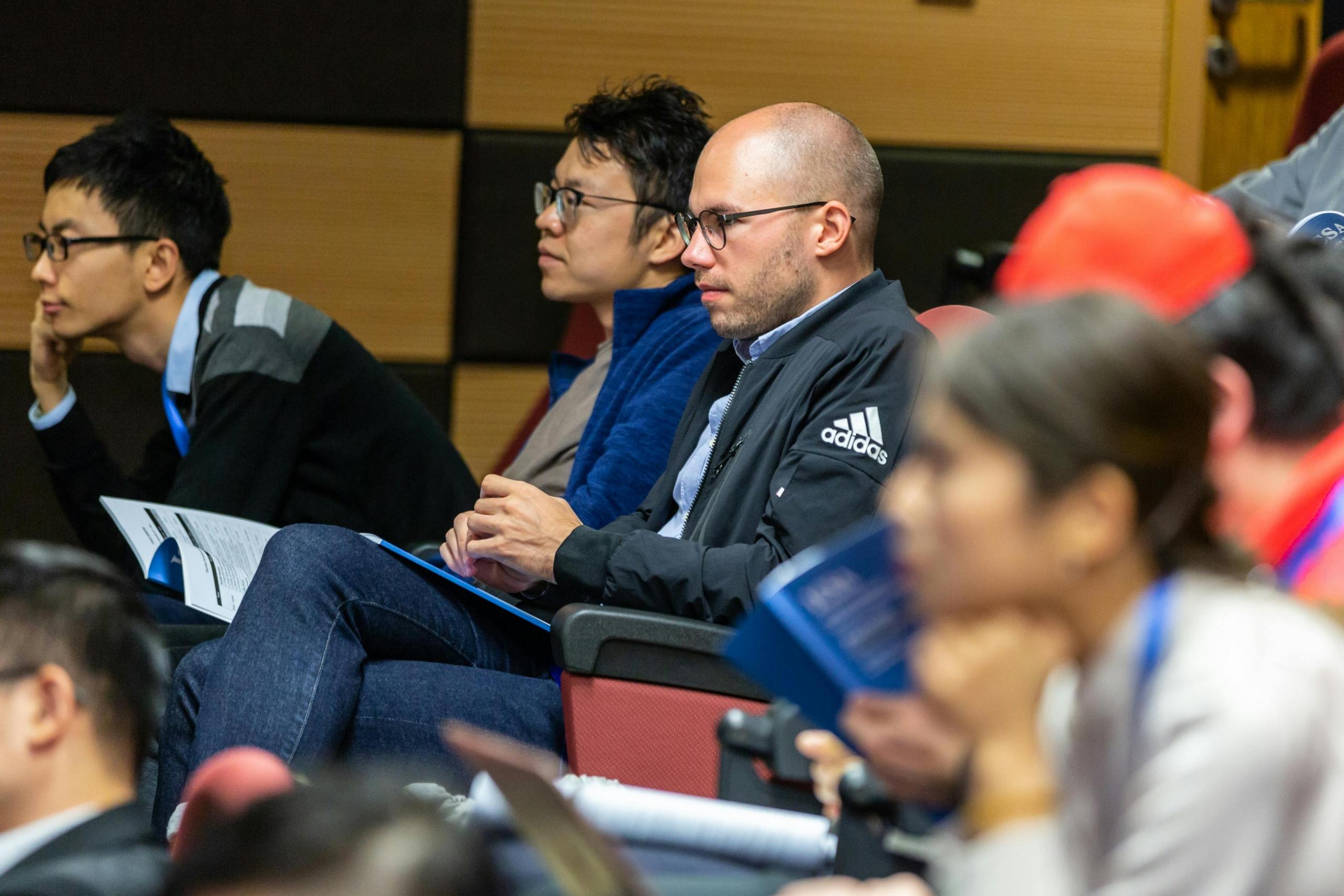 a group of adult students sit in a lecture hall. They appear to be listening to a teacher.