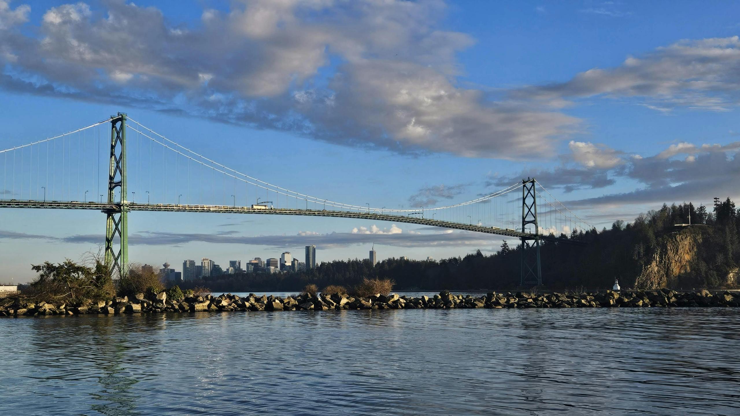 a photo of a bridge crossing the water in Vancouver. The sky above is blue and there are clouds. Green trees can be seen in the distance.