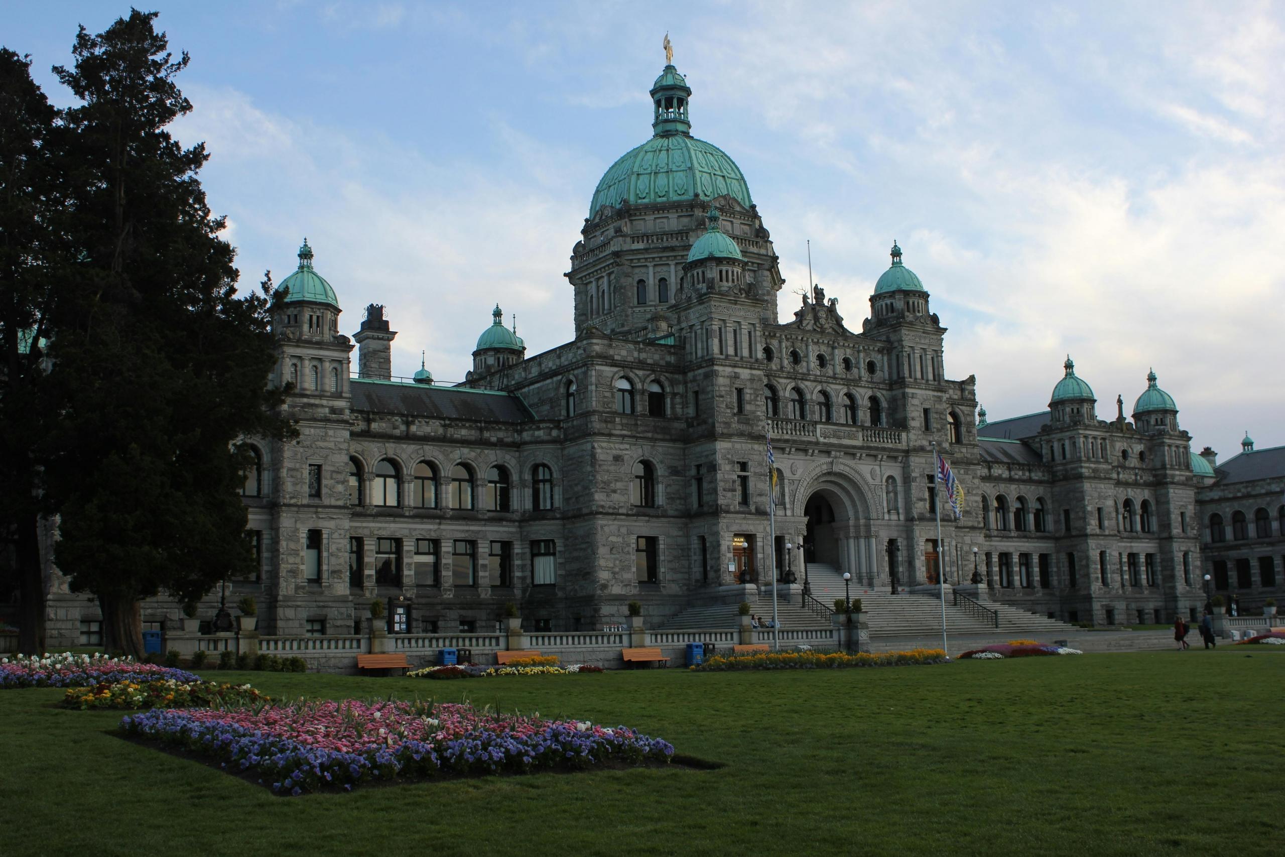 A historic building with a series of green domes stands majestically against a partly cloudy sky. The structure features stonework, numerous arched windows, and a large central entrance. In the foreground, a well-maintained lawn is adorned with colorful flower beds arranged in geometric patterns, adding to the elegance of the scene.