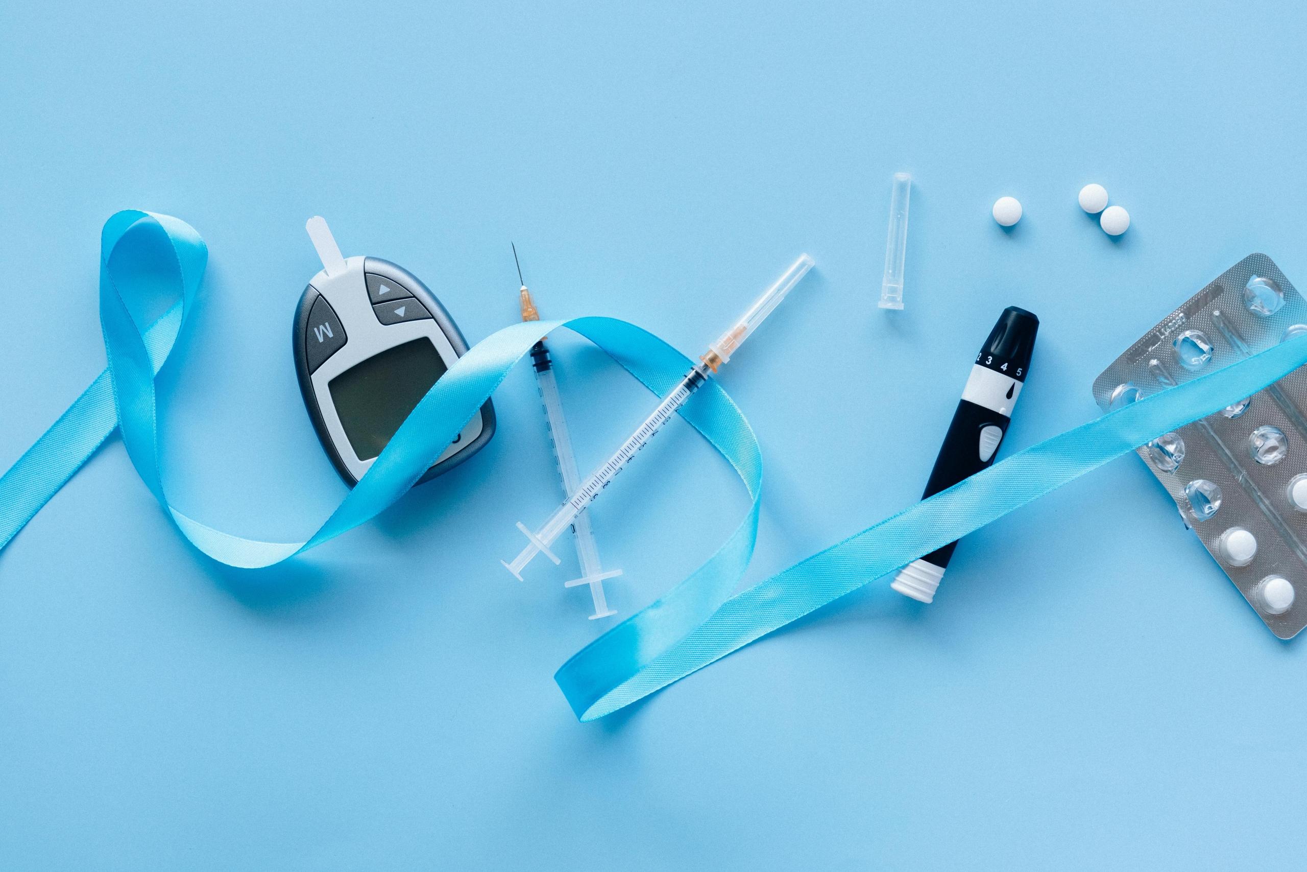 a blood sugar tester, syringes, and pills are on a blue tabletop. A blue ribbon is intertwined.