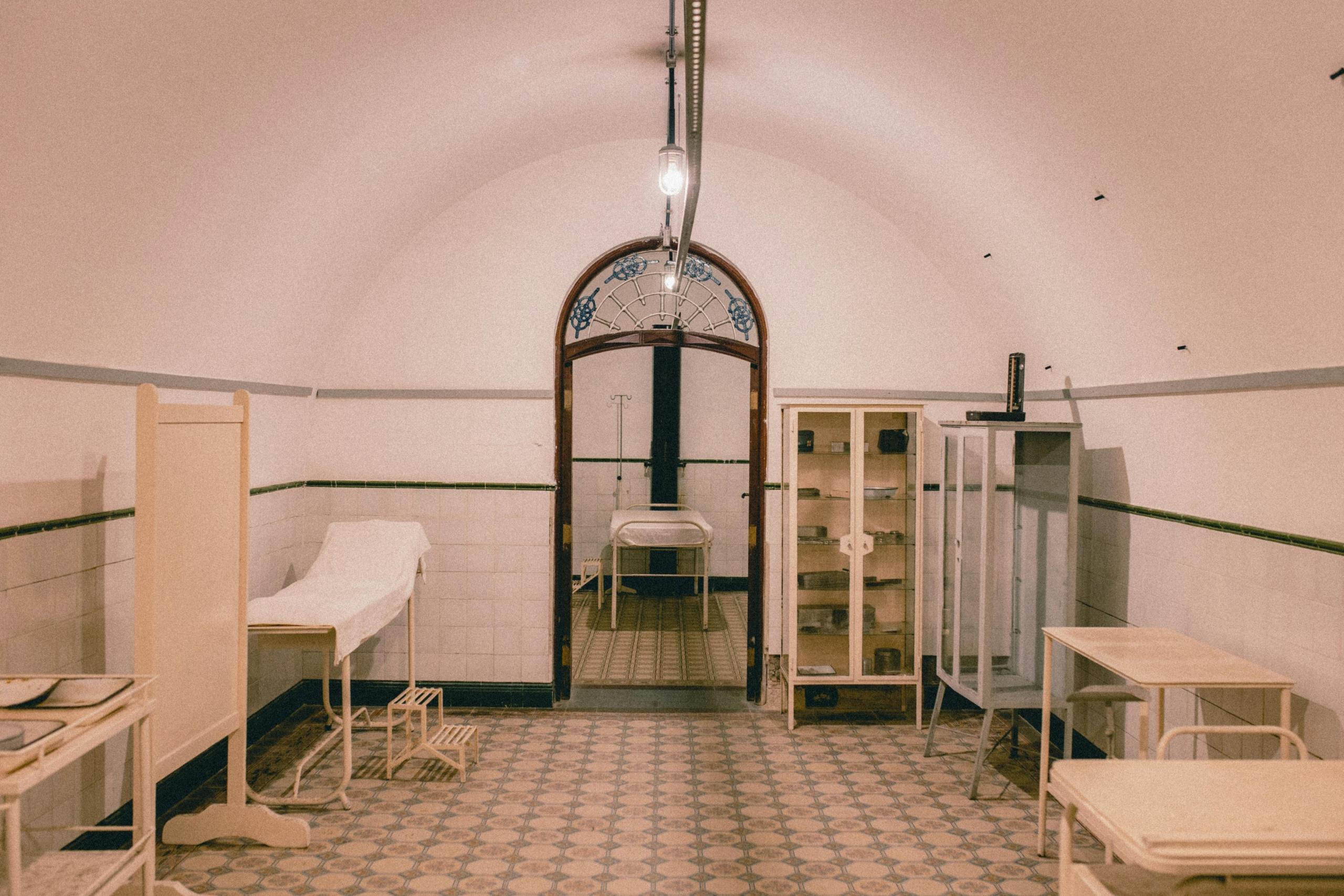 an empty hospital room in an old hospital. A bed can be seen one one side and a doorway leads to another room.