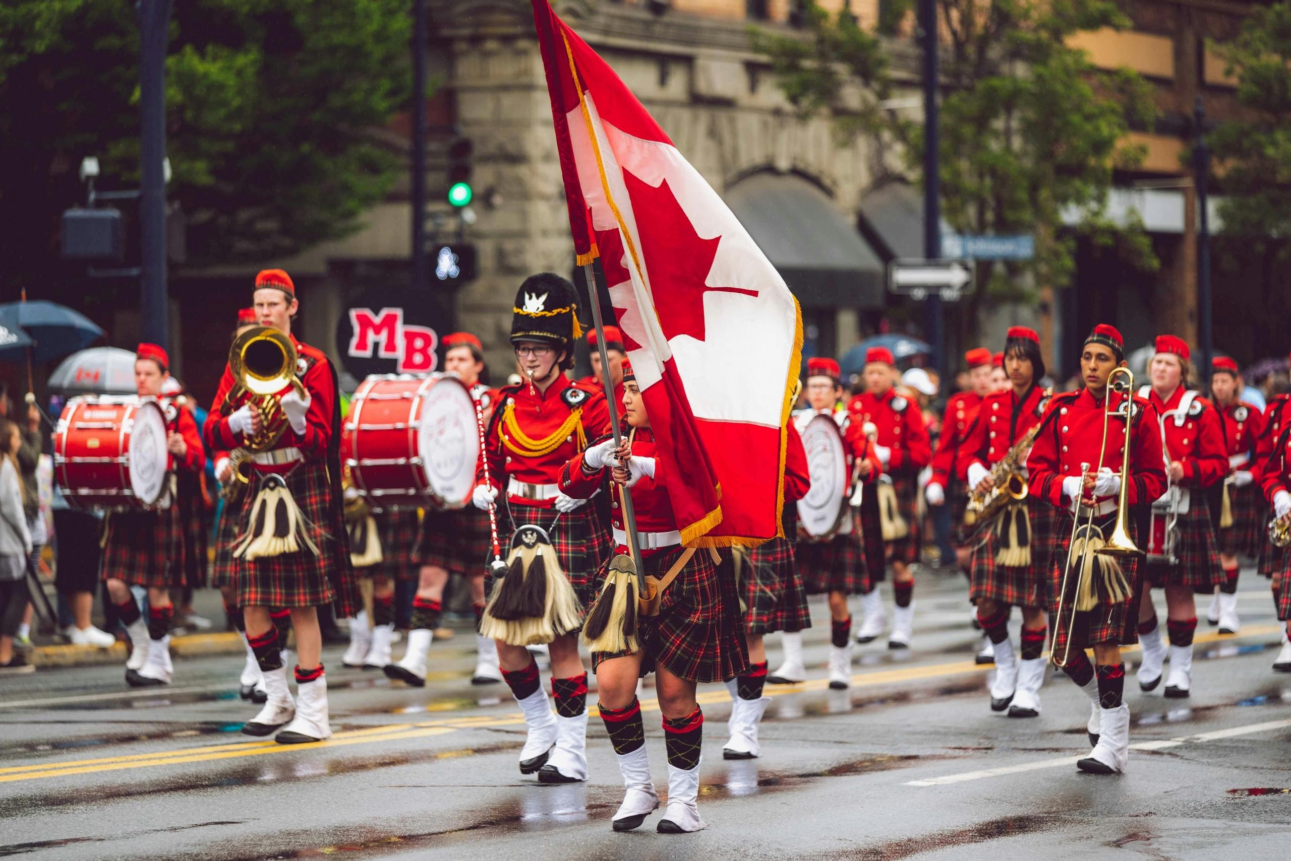 A parade featuring Canadian flag bearers and a marching band in traditional red uniforms and kilts, performing on a city street.