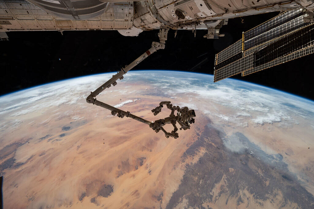 Canadarm2 robotic arm extending from the International Space Station over Earth, with the Dextre robotic manipulator attached to its end and one of the station's solar arrays visible against the blackness of space.