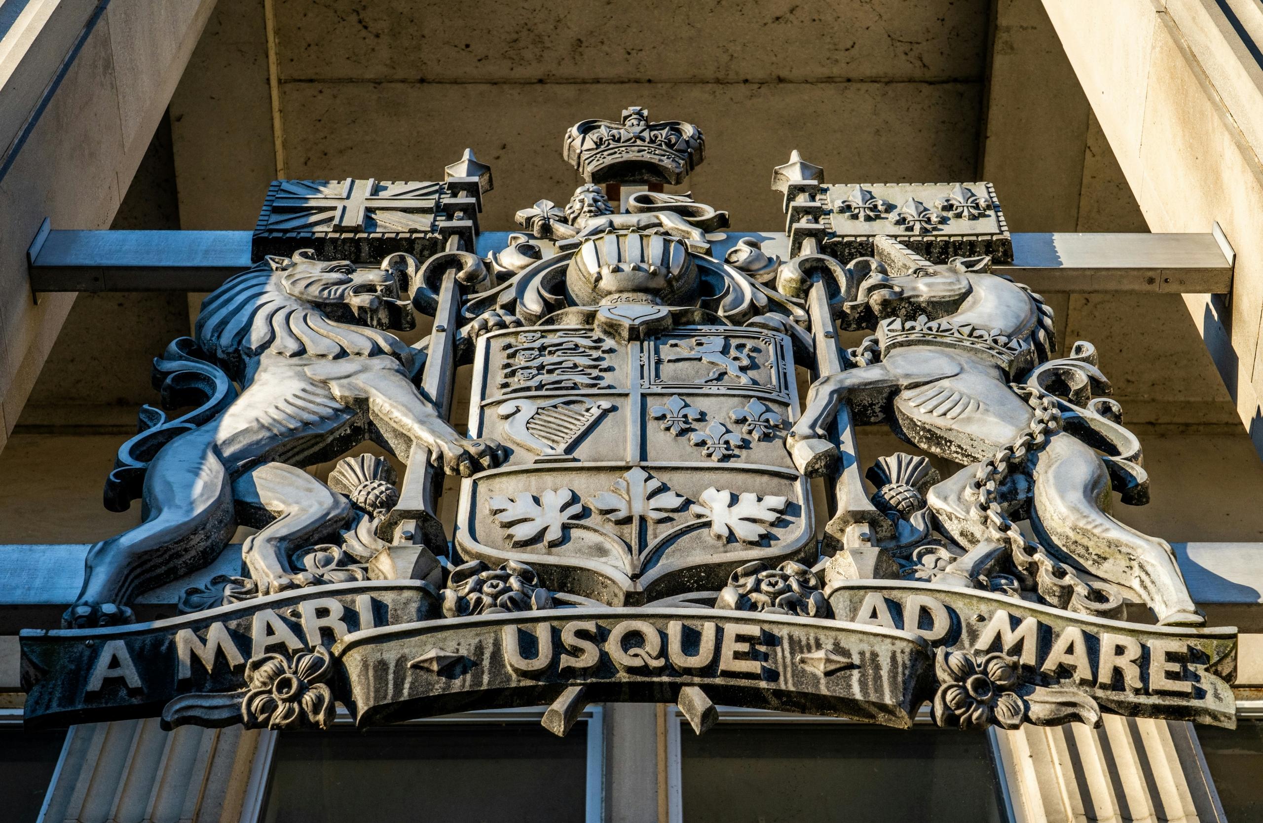 A close-up view of the Canadian Coat of Arms carved on a government building, featuring the lion, unicorn, and national symbols.
