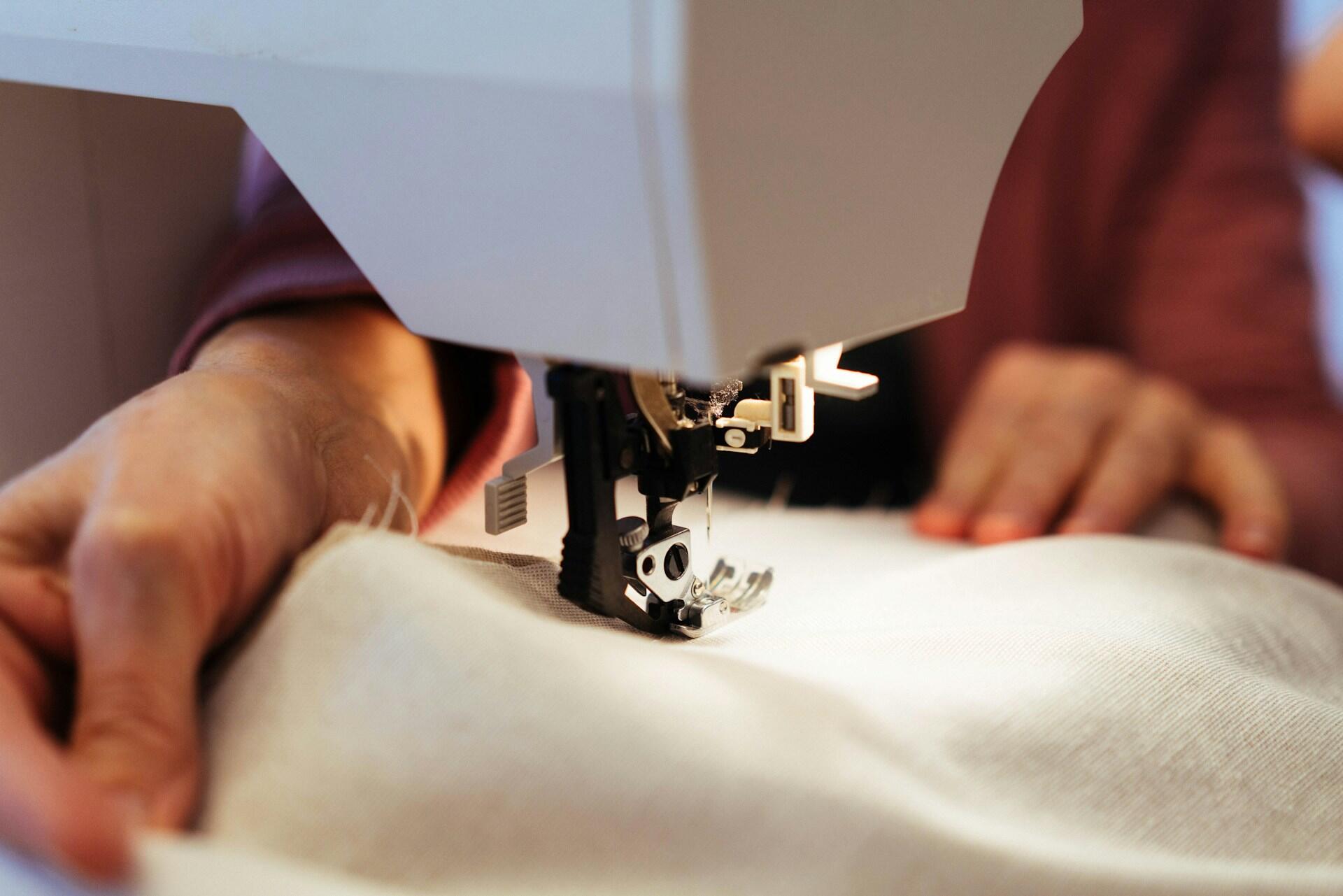 Close-up of hands guiding beige fabric through a sewing machine during stitching, with part of the machine and needle in focus.