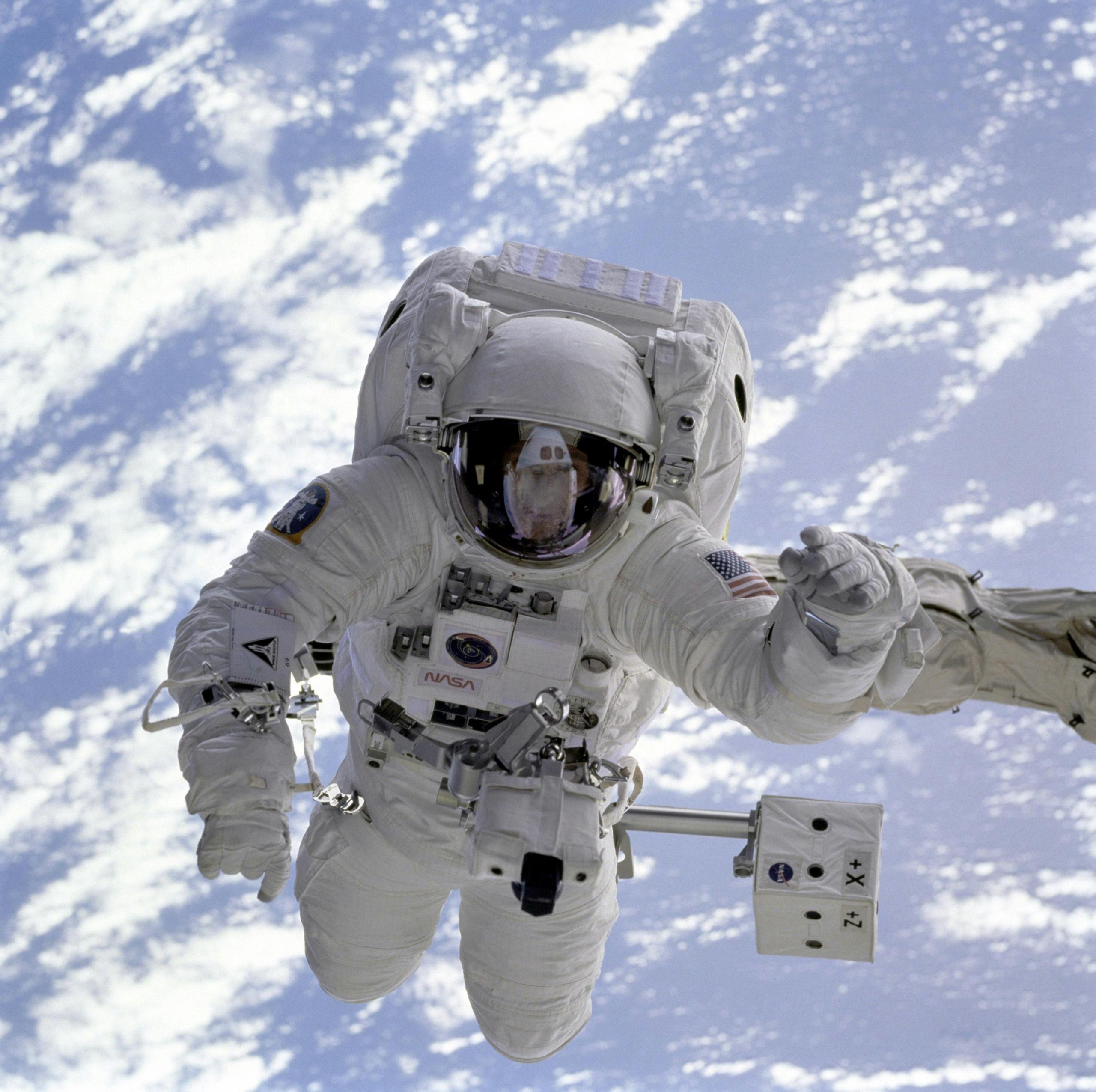 A NASA astronot floats in space above earth. The astronaut wears a white spacesuit and reflective helmet.