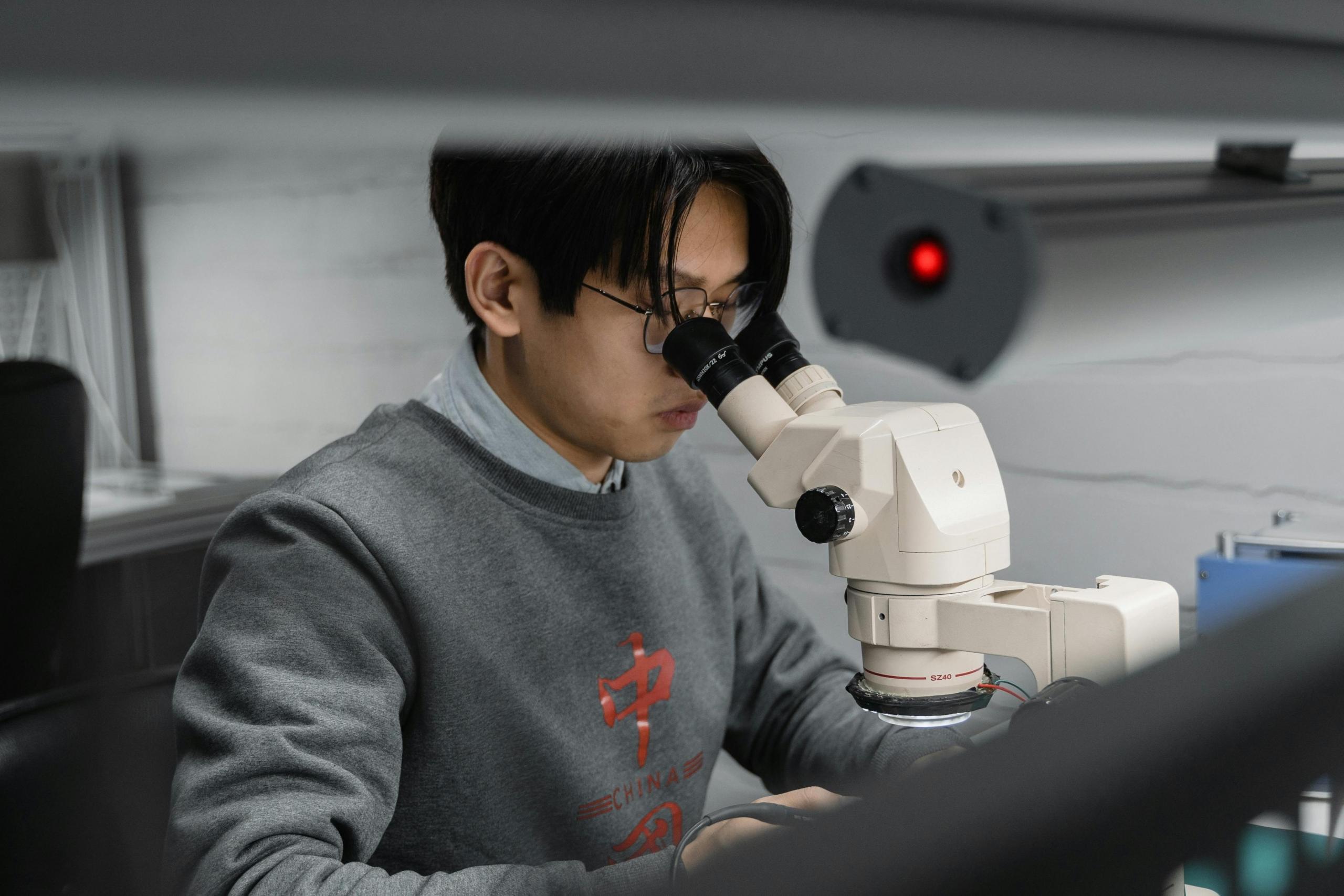 A male student in a gray sweatshirt looks into a powerful microscope.