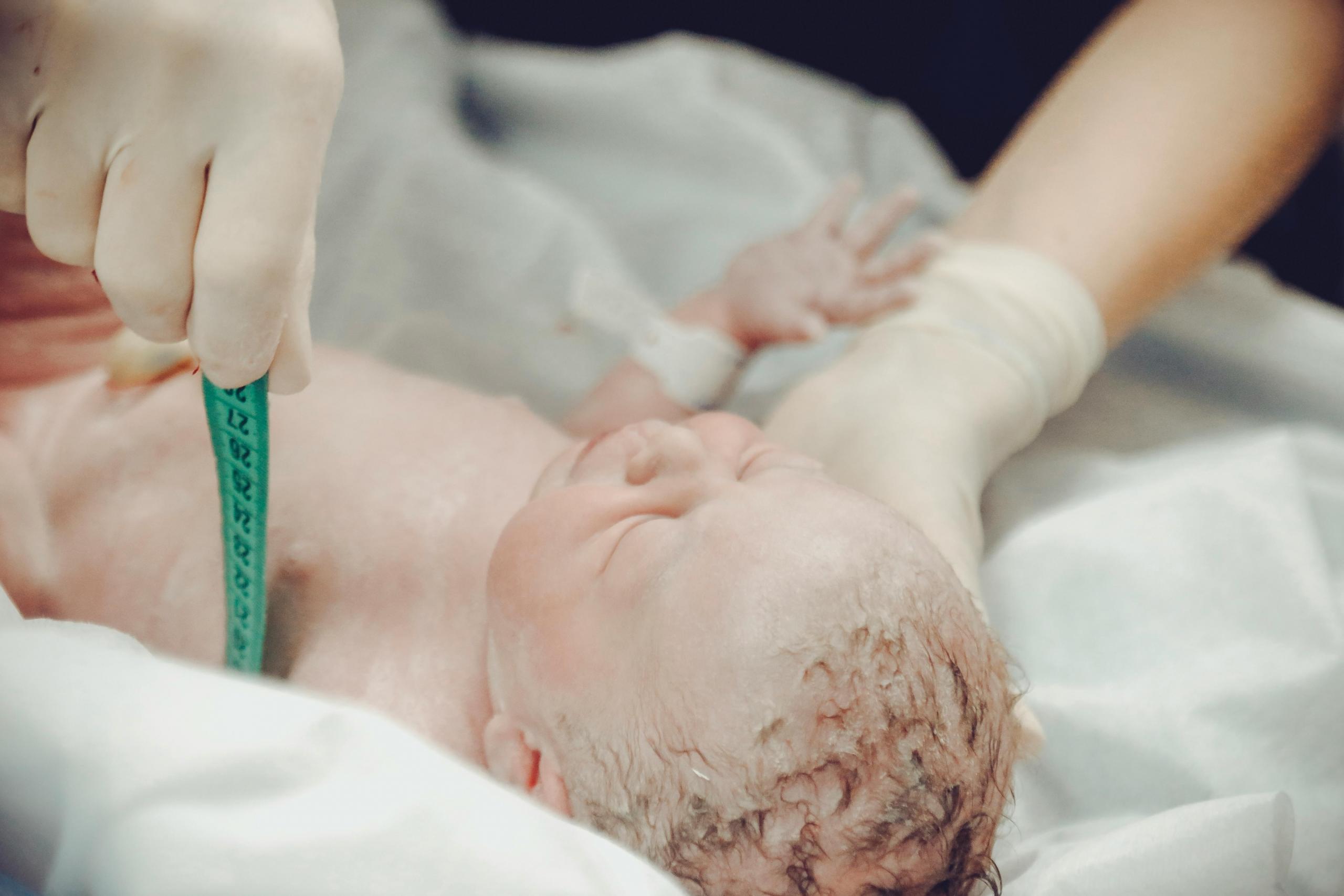 a newborn baby is measured by medical staff. A nurse holds a green tape measure to check the baby's size. The baby has their hand outstretched and eyes closed. 