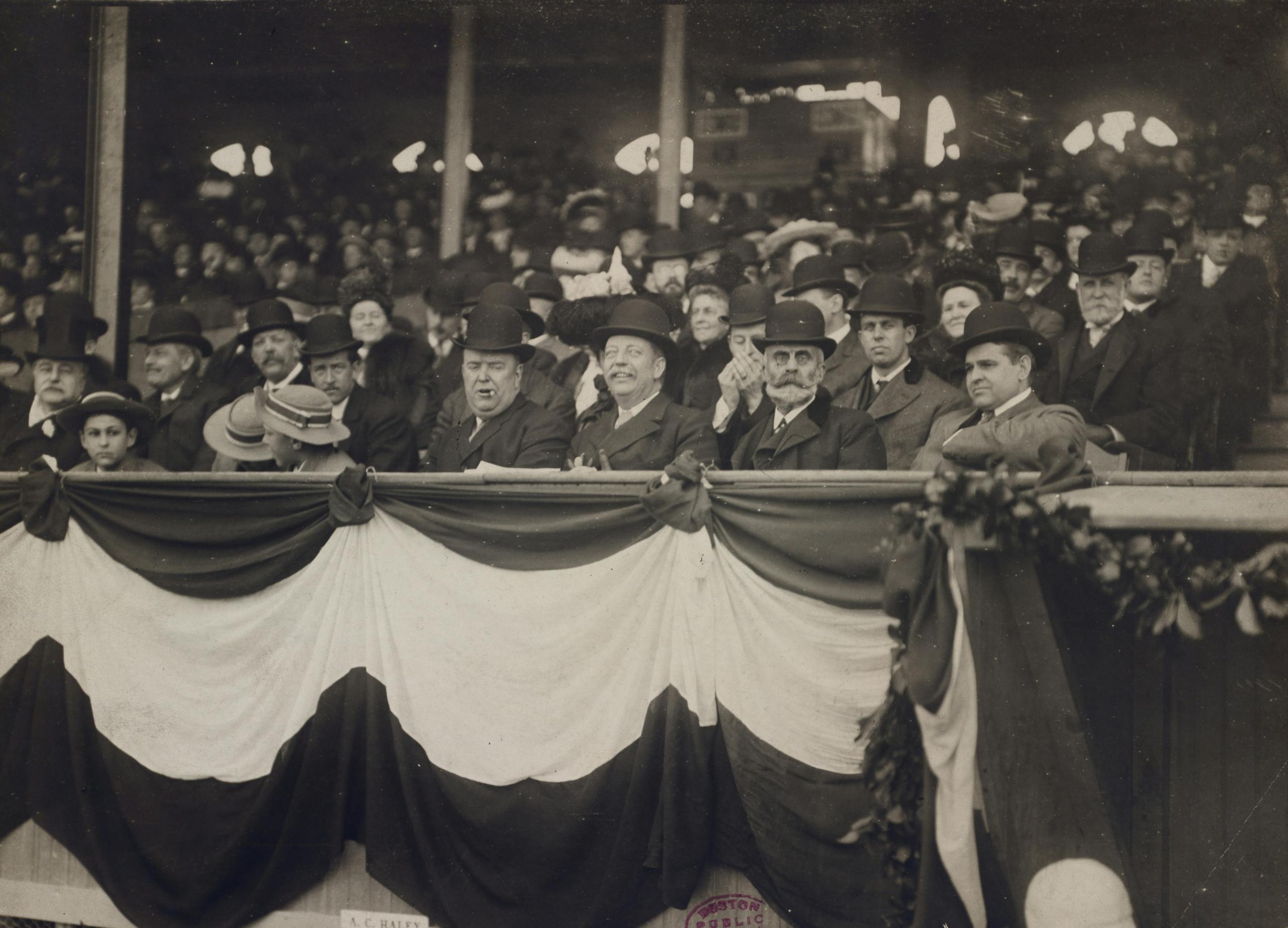 A black-and-white photograph of spectators in the early 1900s, most top hats made from beaver fur felt, seated in a grandstand at a formal event.