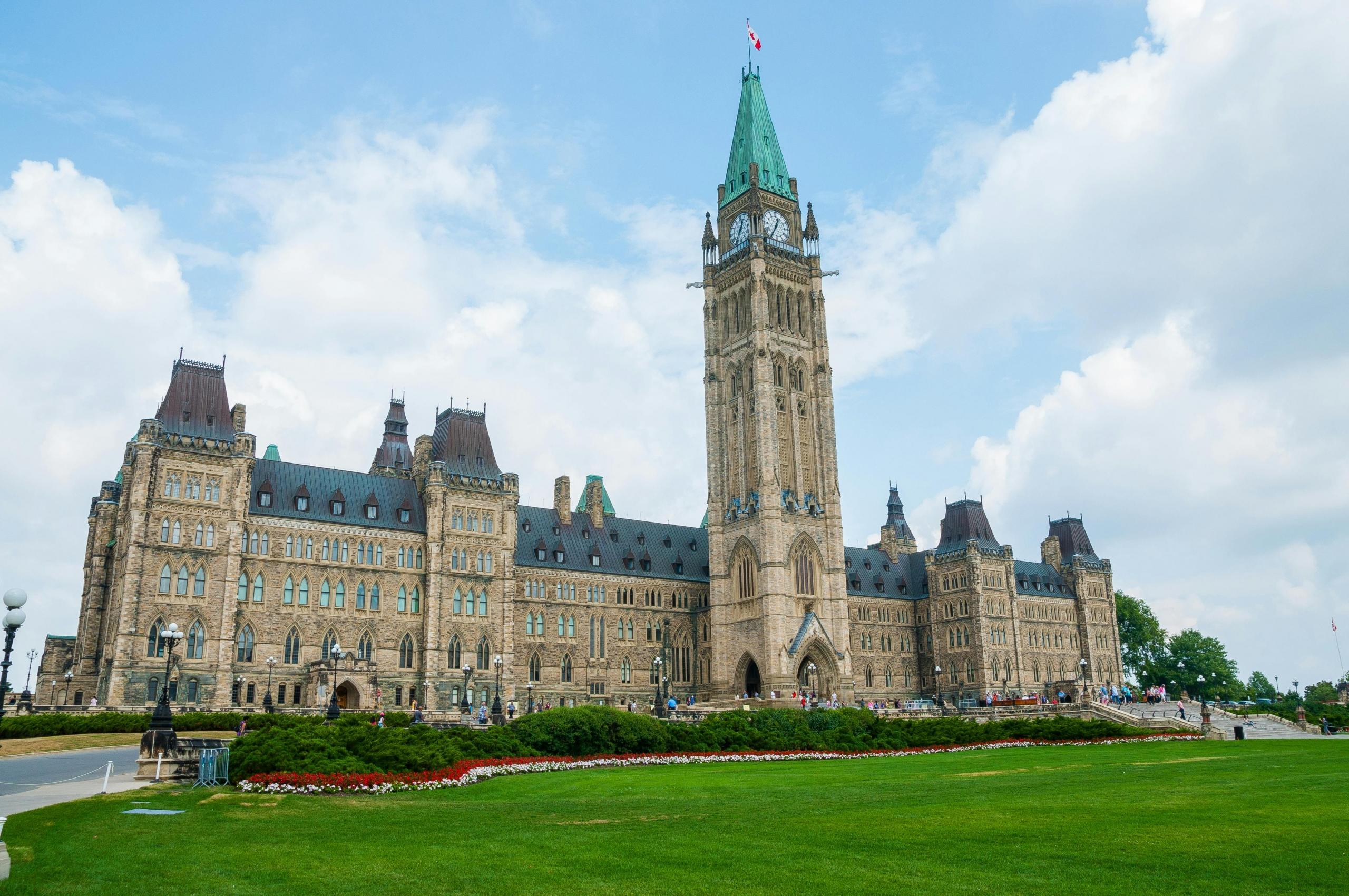 The Canadian Parliament Building in Ottawa on a sunny day, showcasing its Gothic Revival architecture with the Peace Tower at the center and green lawns in the foreground.