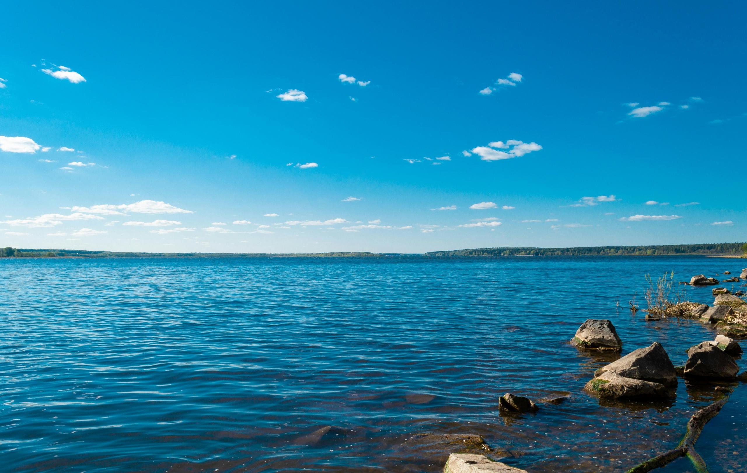 a lake is empty and reflects the sky. Land with trees can be seen in the distance, and rocks are at the forefront of the picture.