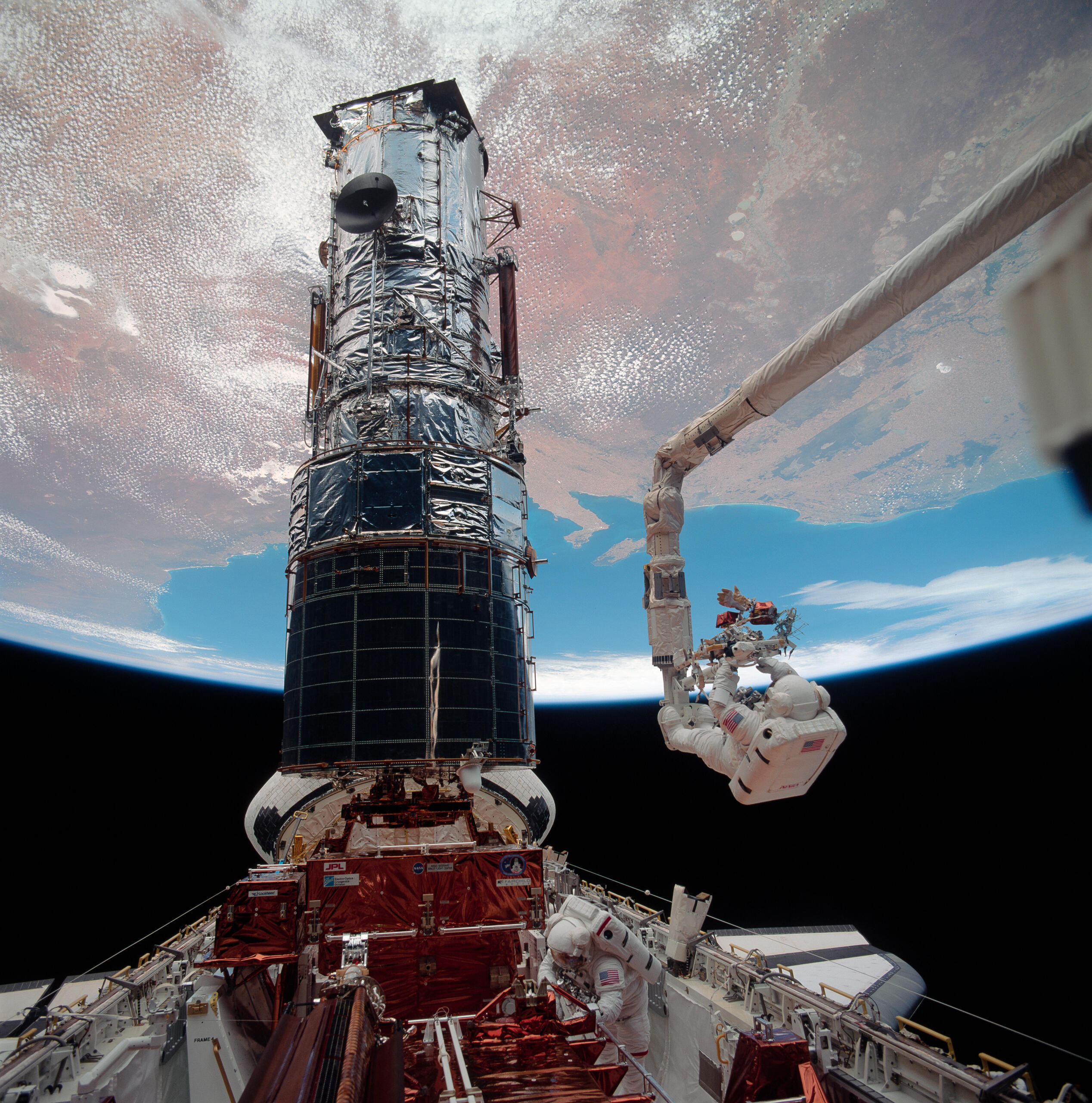 Astronauts performing a spacewalk during the Hubble Space Telescope servicing mission, with one astronaut attached to the Space Shuttle's Canadarm while working on the telescope above Earth's surface.