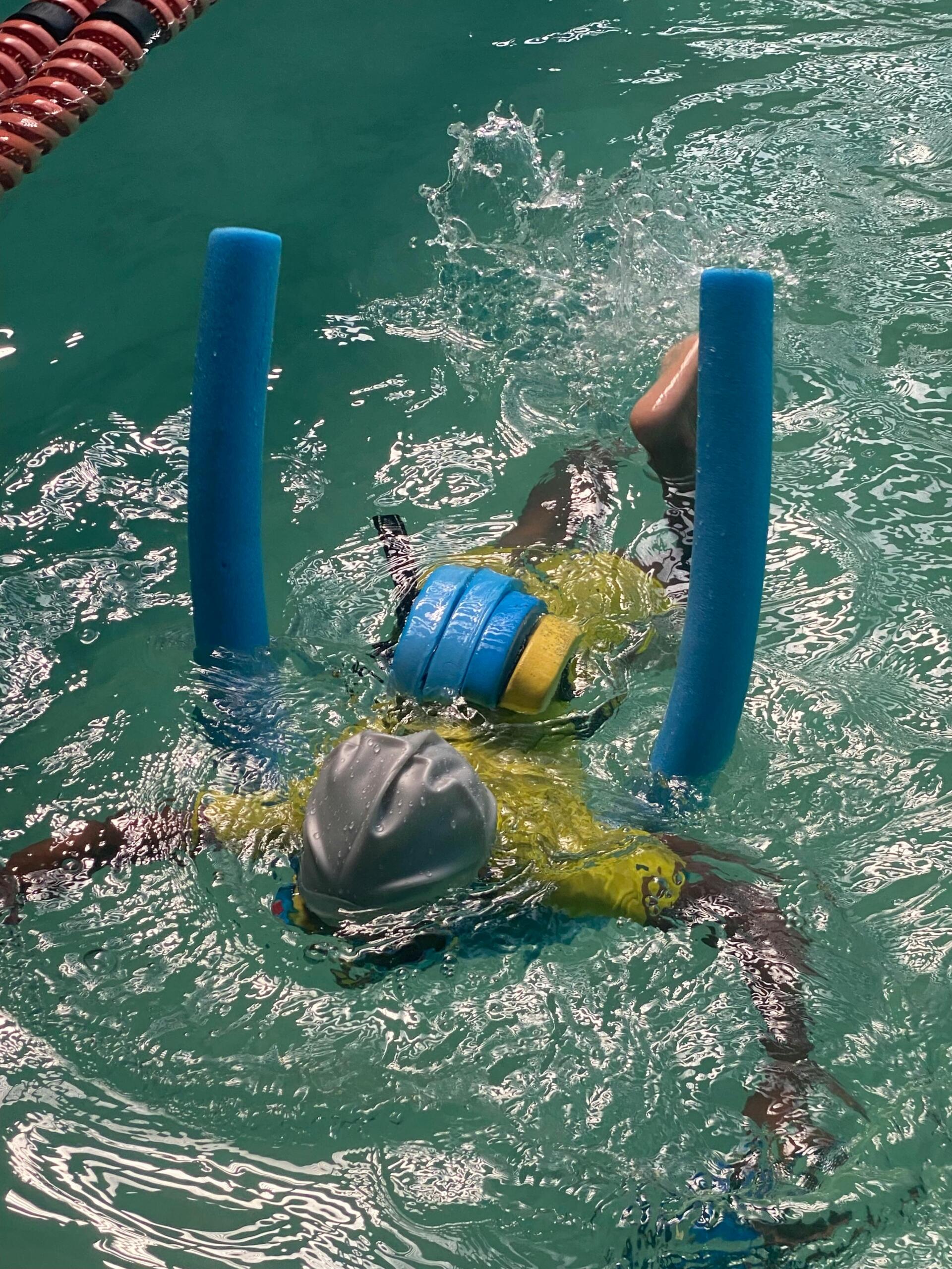 A child swims in an indoor pool using multiple flotation devices, including arm floats and a kickboard, while practicing safely in shallow water.