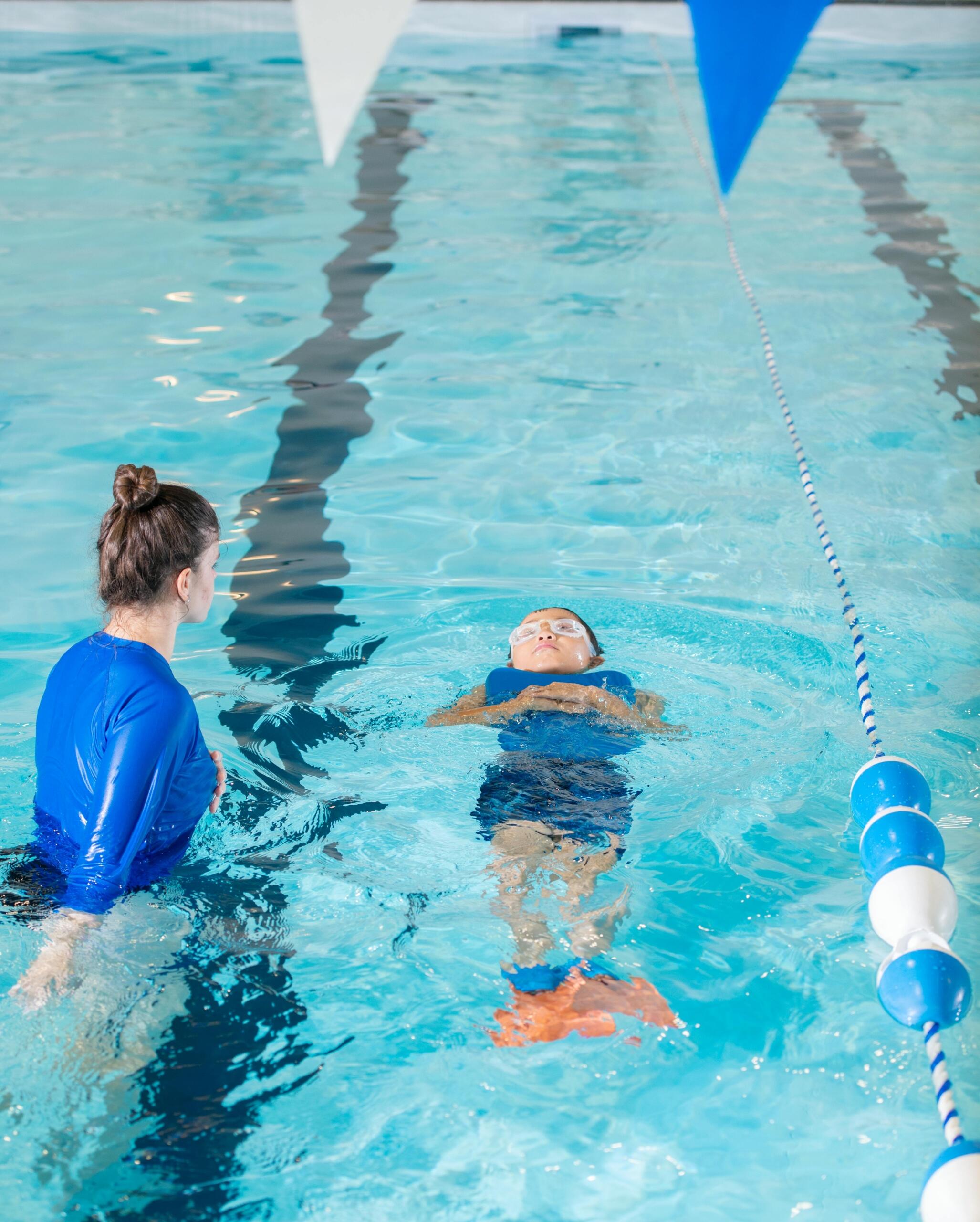 A young boy practices swimming in a pool while a swim instructor guides his movement and helps him float with confidence.