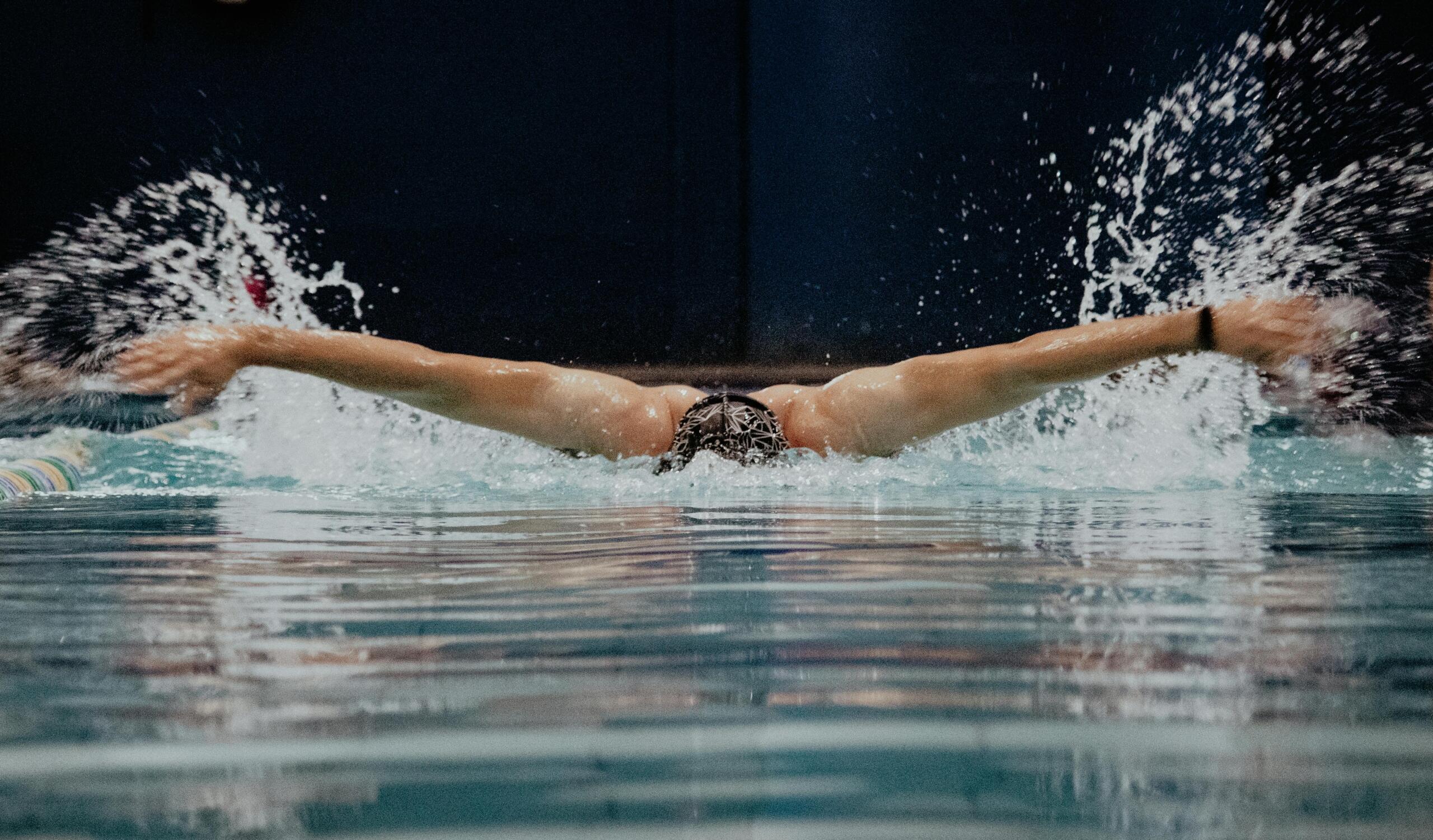 Swimmer performing butterfly stroke in an indoor pool, arms extended mid-stroke with water splashing