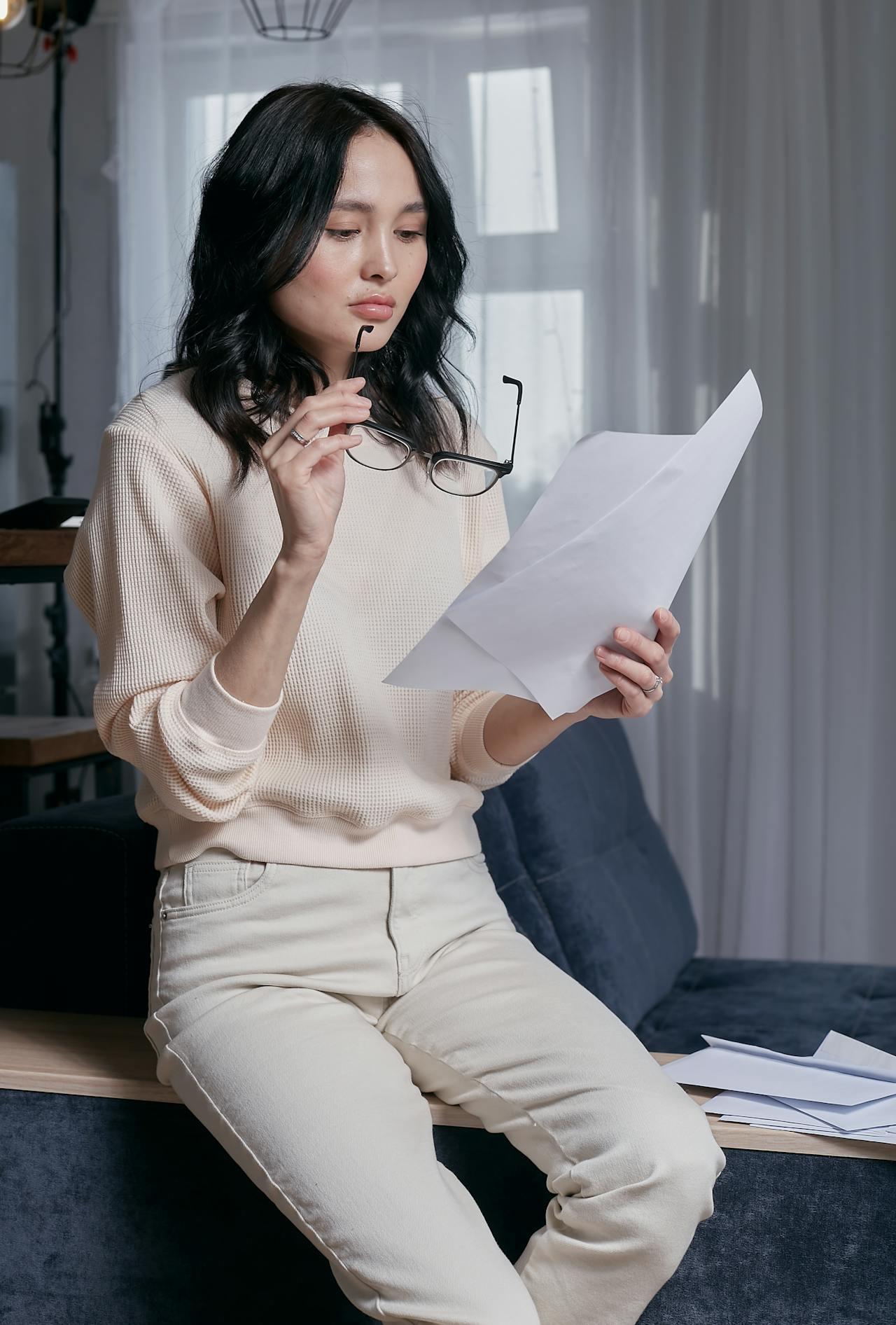 A women leaning against a desk with a paper in one hand and glasses in the other.