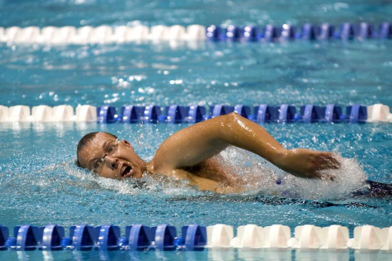 A swimmer in an indoor pool executes a freestyle stroke, creating splashes amid blue water and lane dividers.