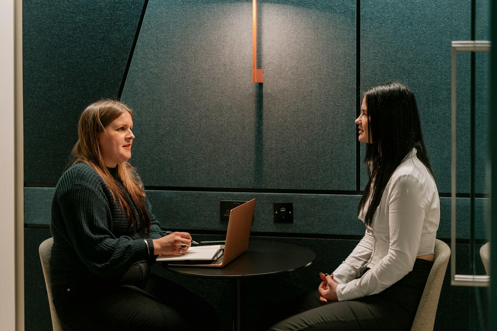 Two individuals in a meeting room with one using a laptop on a round table.