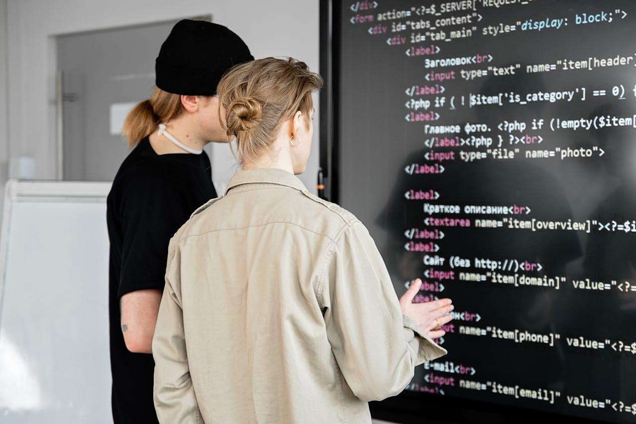 Two individuals stand before a large screen displaying programming code, engaged in a discussion about website features and design.