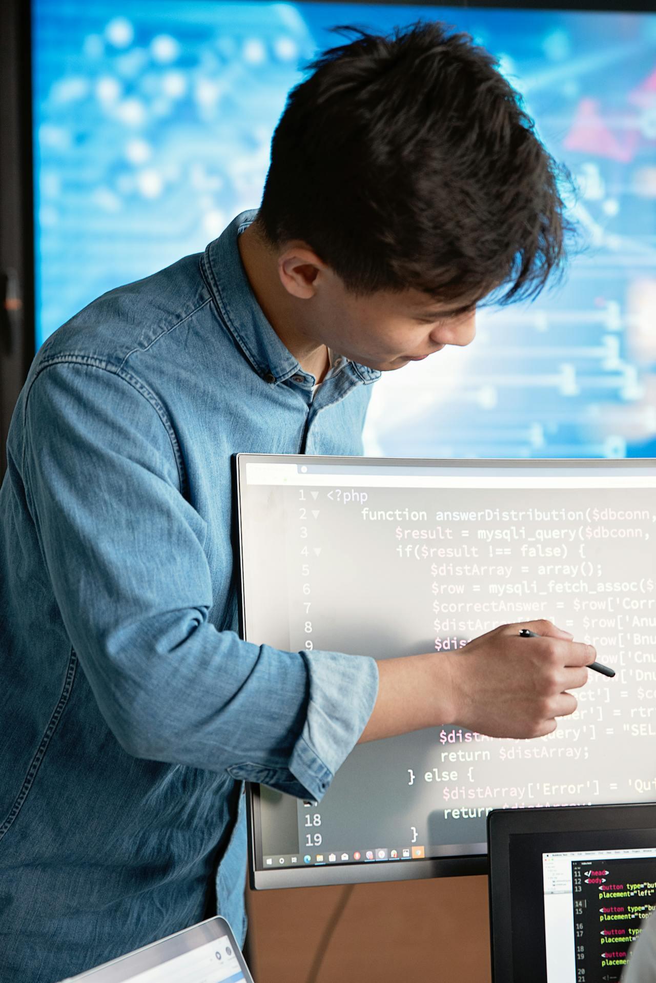 A person in a denim shirt points at code displayed on a computer screen, with programming content visible in the background.