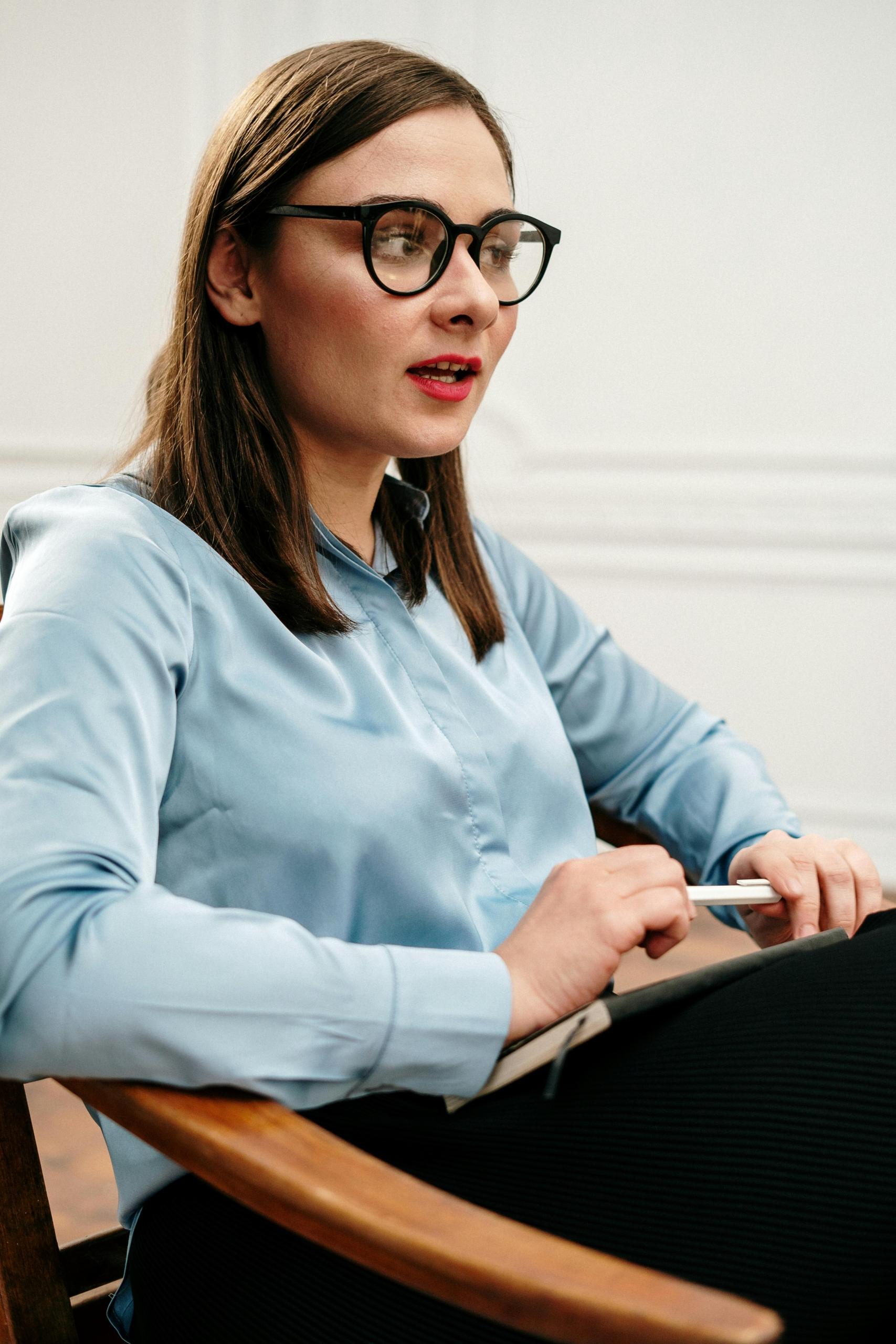 a female professional sits in a chair. She wears glasses and a blue shirt and listens to another person.