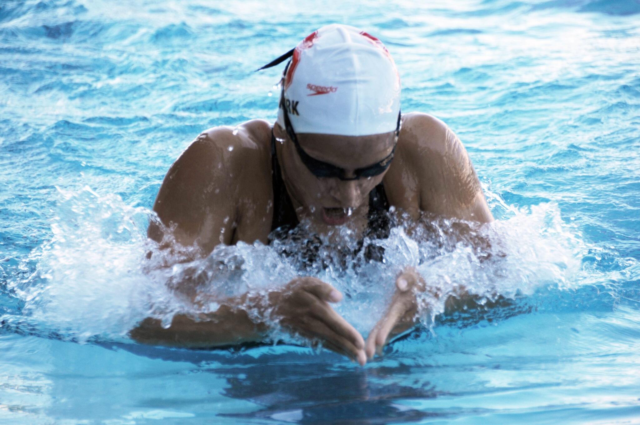 A swimmer performs a breaststroke in a pool, creating splashes of water around their arms and legs against a backdrop of blue water.