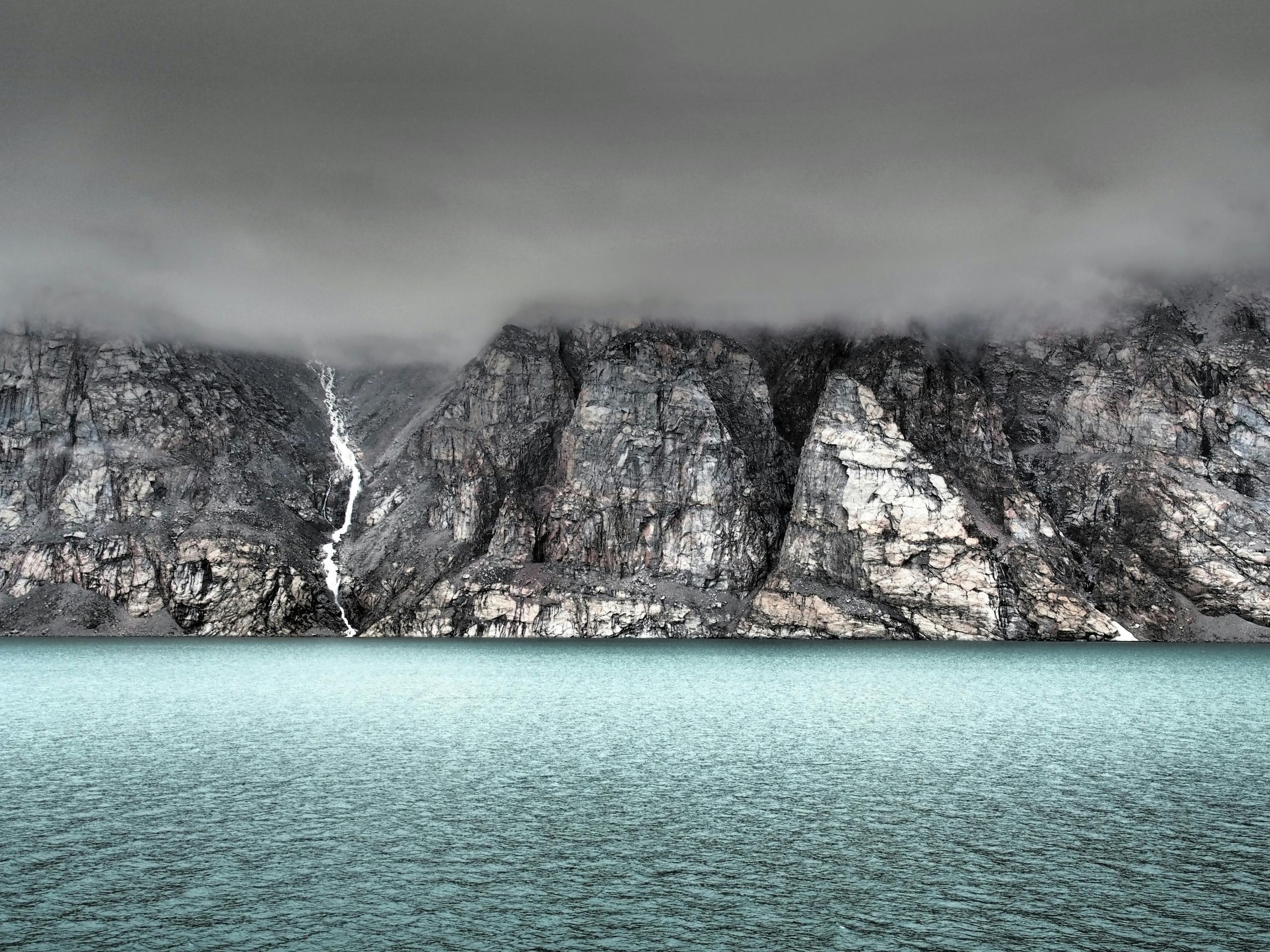 Rugged cliffs of Baffin Island with mist overhead and glacial streams flowing into the blue Arctic waters.
