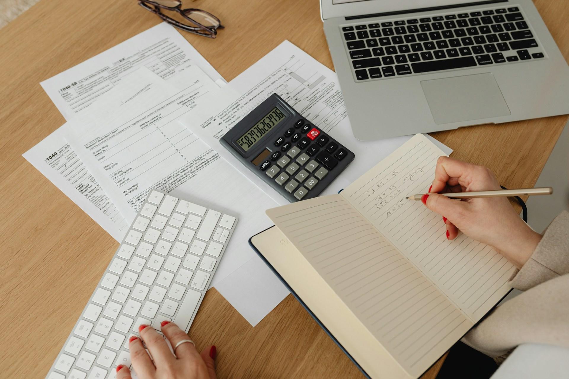 Person calculating finances with laptop, papers, calculator, and notepad on desk.