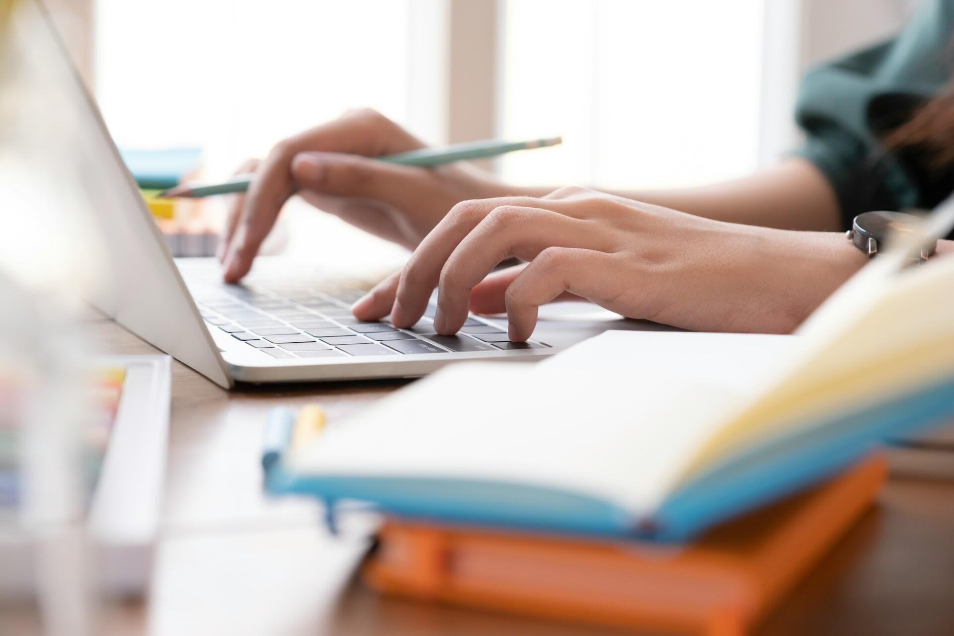 Hands typing on a laptop keyboard with a pencil in one hand and an open notebook on the desk nearby.