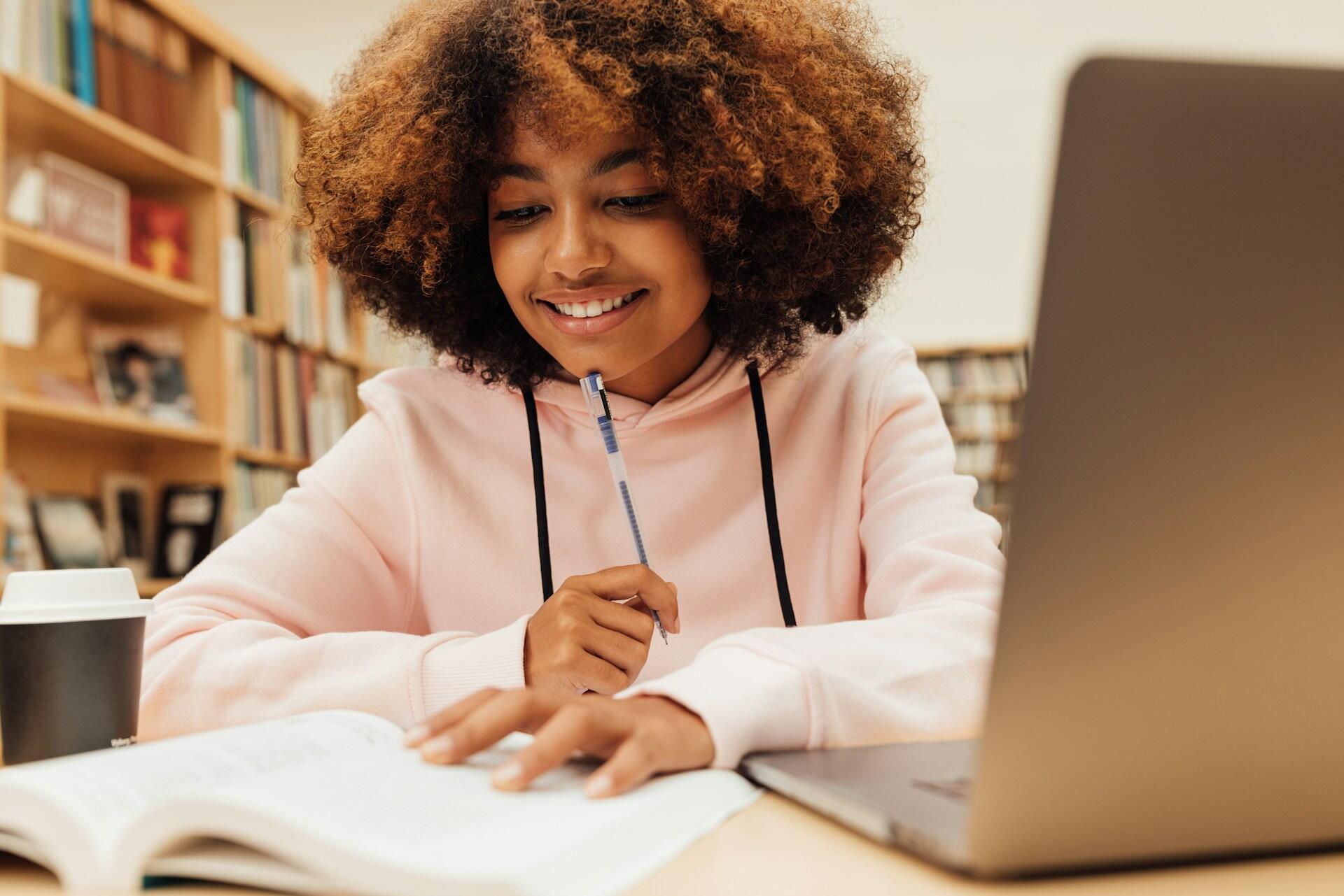 Person in a pink hoodie studying with a book and laptop in a library, holding a pen near their chin and a coffee cup nearby.