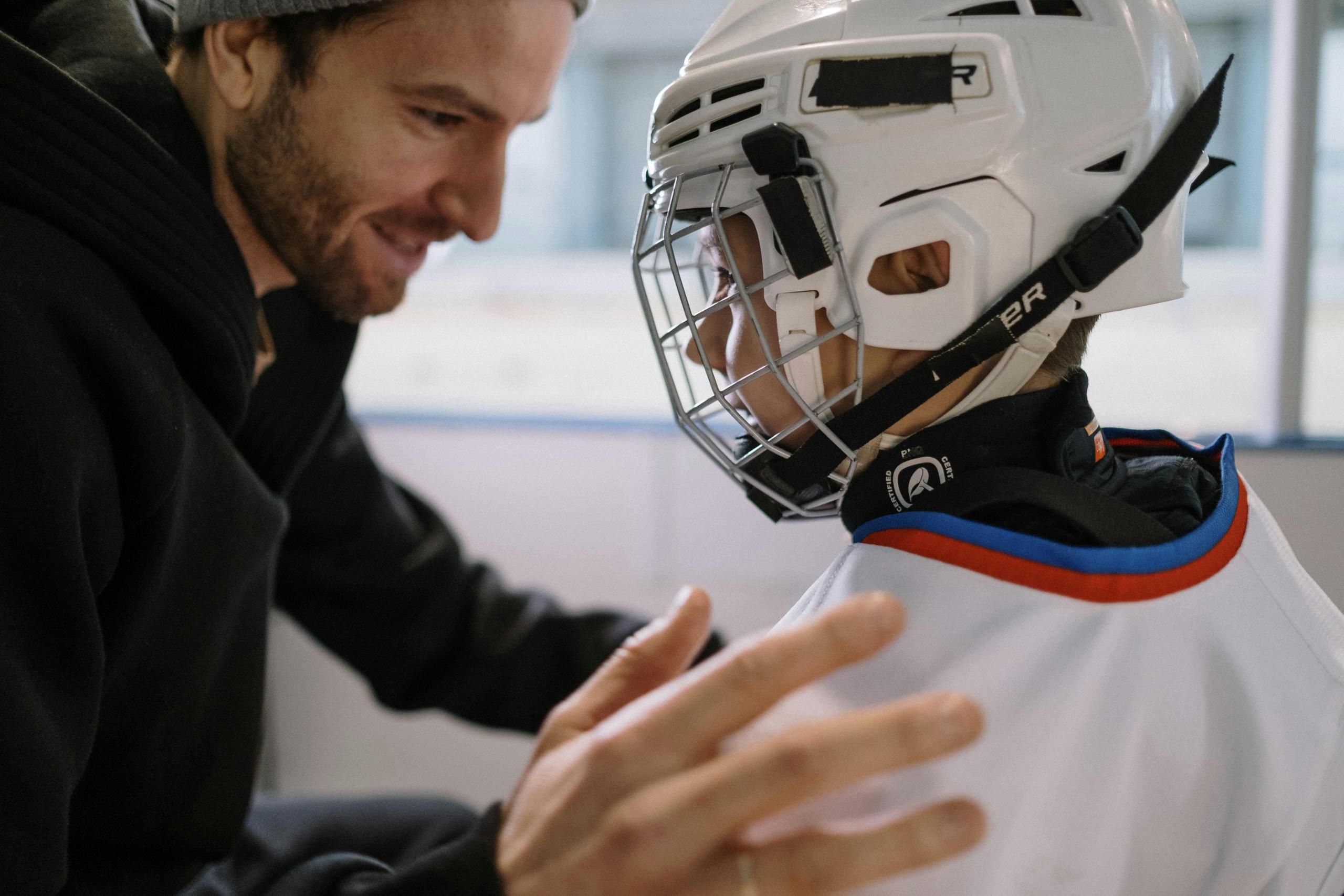 a father looks lovingly at his son, who wears a hockey helmet and uniform.
