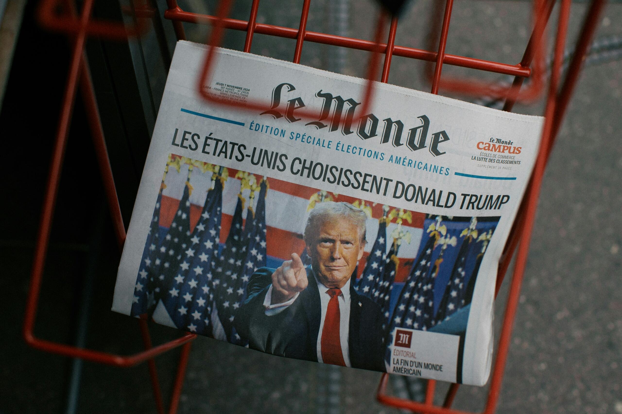 A close-up of a newspaper titled 'Le Monde' featuring an article about Donald Trump, set against a backdrop of American flags.