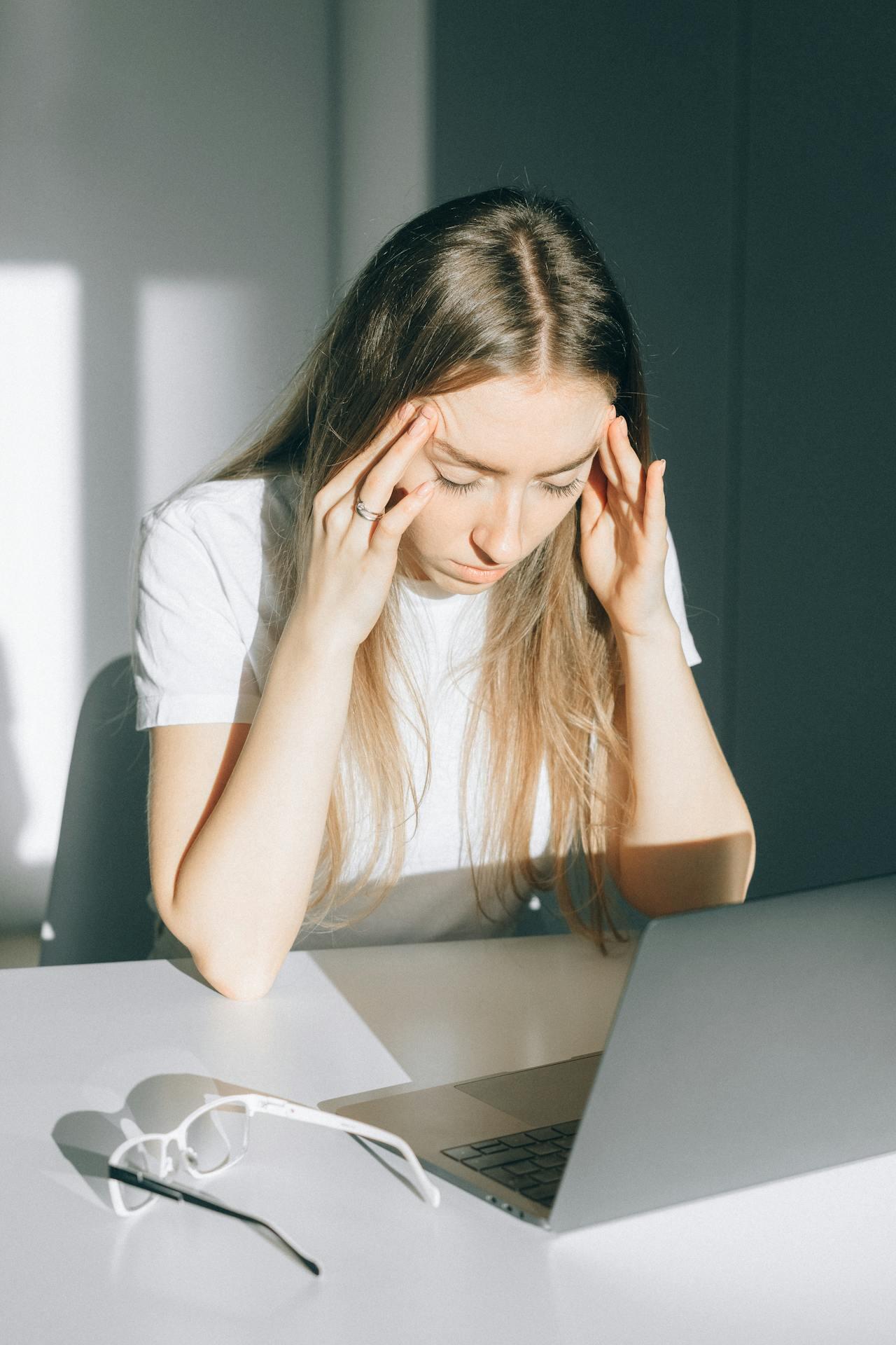 Woman with long hair in white shirt sitting at a desk holding her head, with a laptop and glasses on the table in bright lighting.
