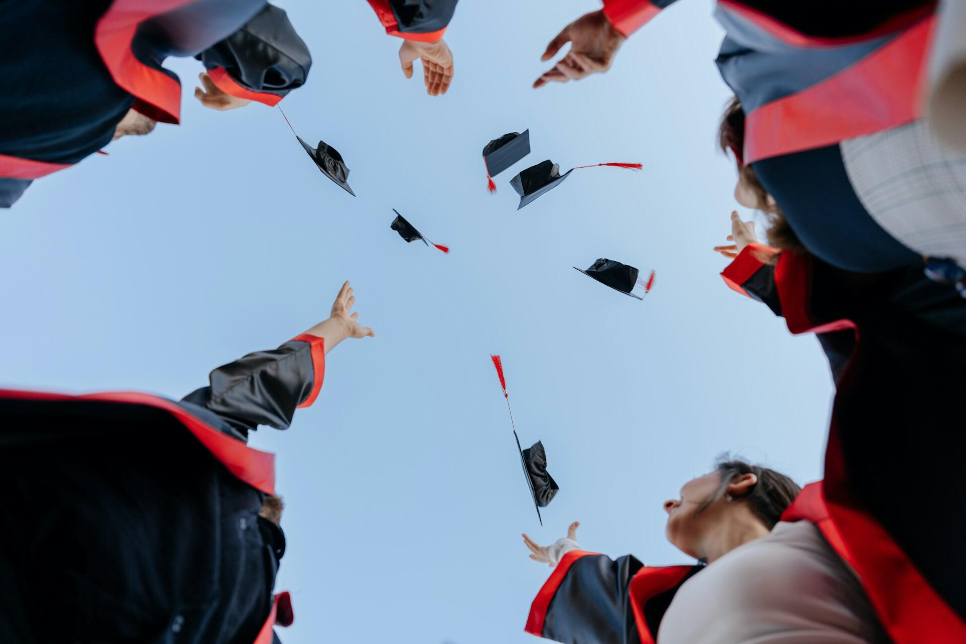 Graduates in black and red gowns throw graduation caps into a clear blue sky in celebration.