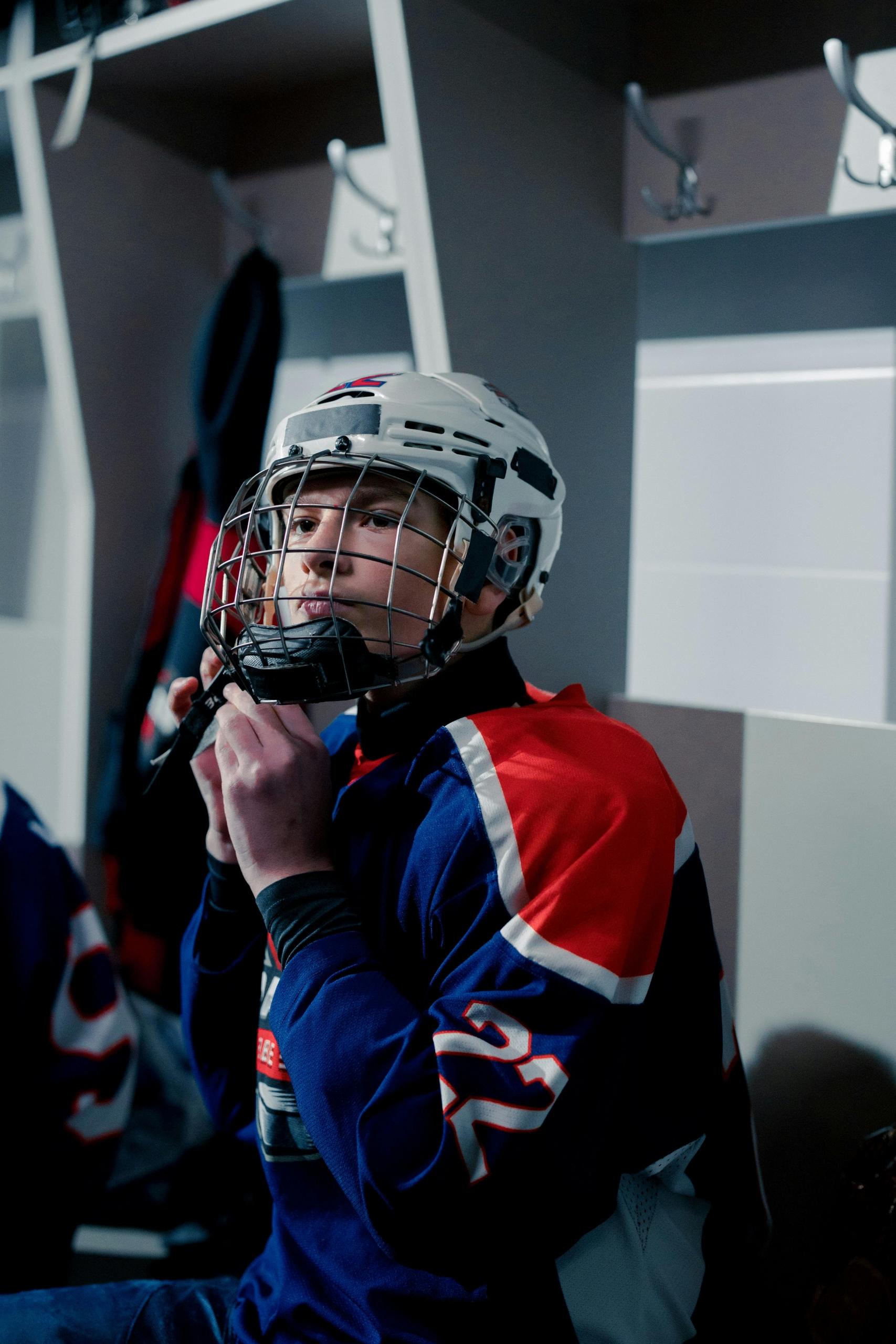 a boy in a stadium change room adjusts his hockey helmet.