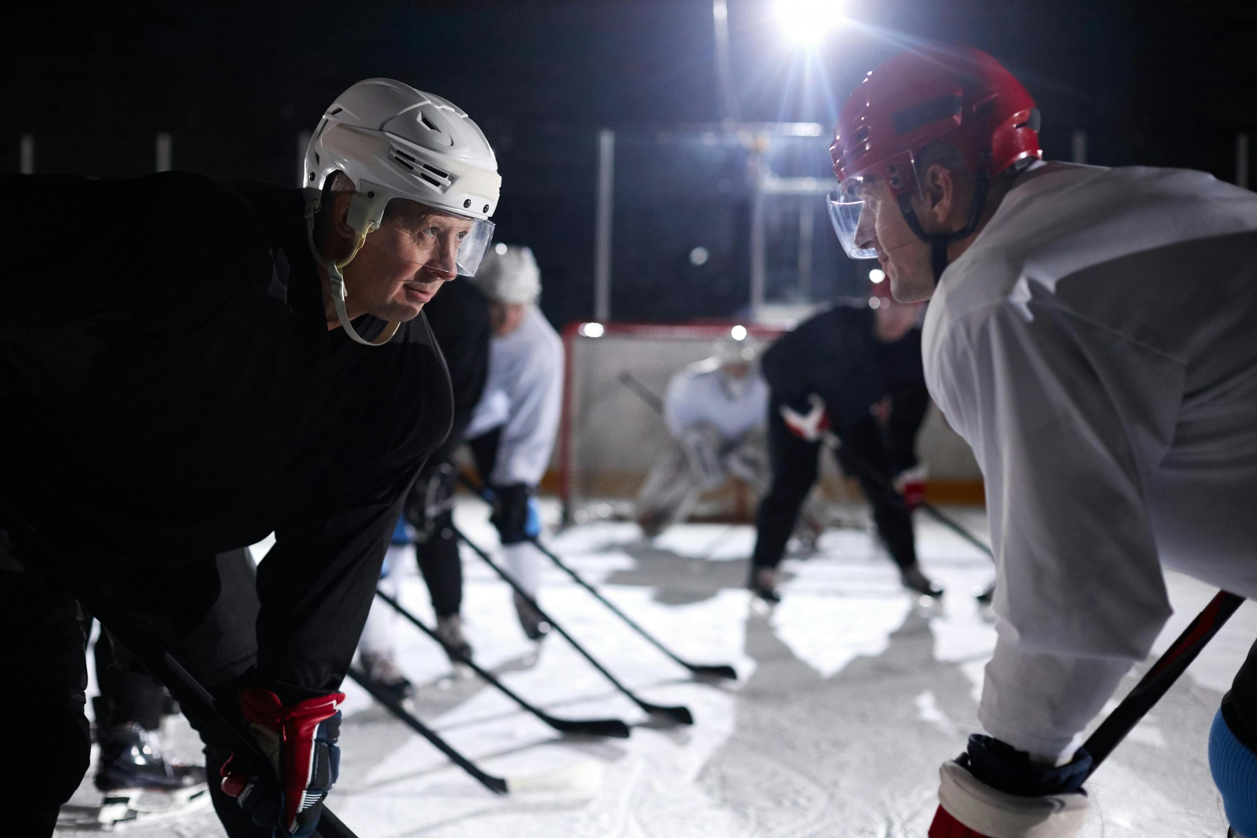 Two men face each other at a hockey game. One man wears a white helmet, the other wears a red helmet. They are on an ice rink, and other players can be seen in the background.