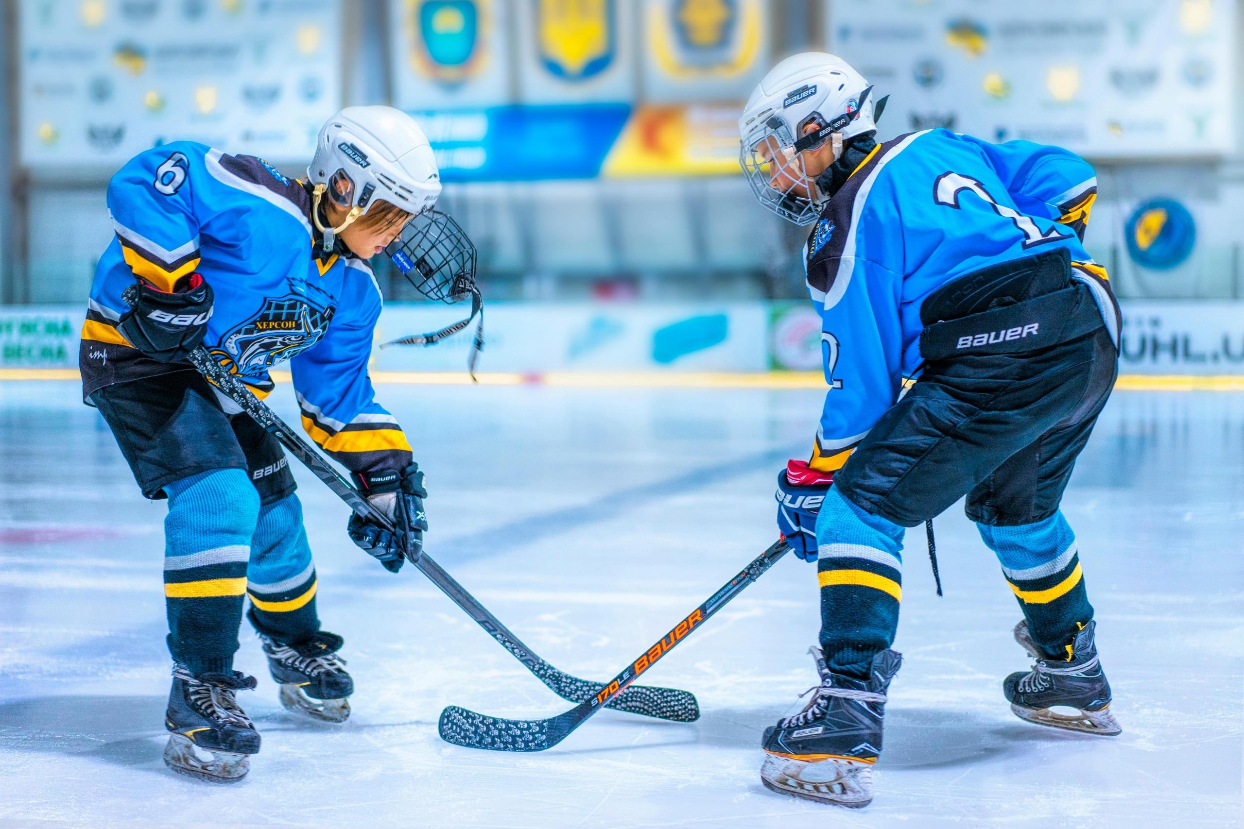 Two young people wear blue hockey uniforms and face off at a hockey rink. They hold hockey sticks.