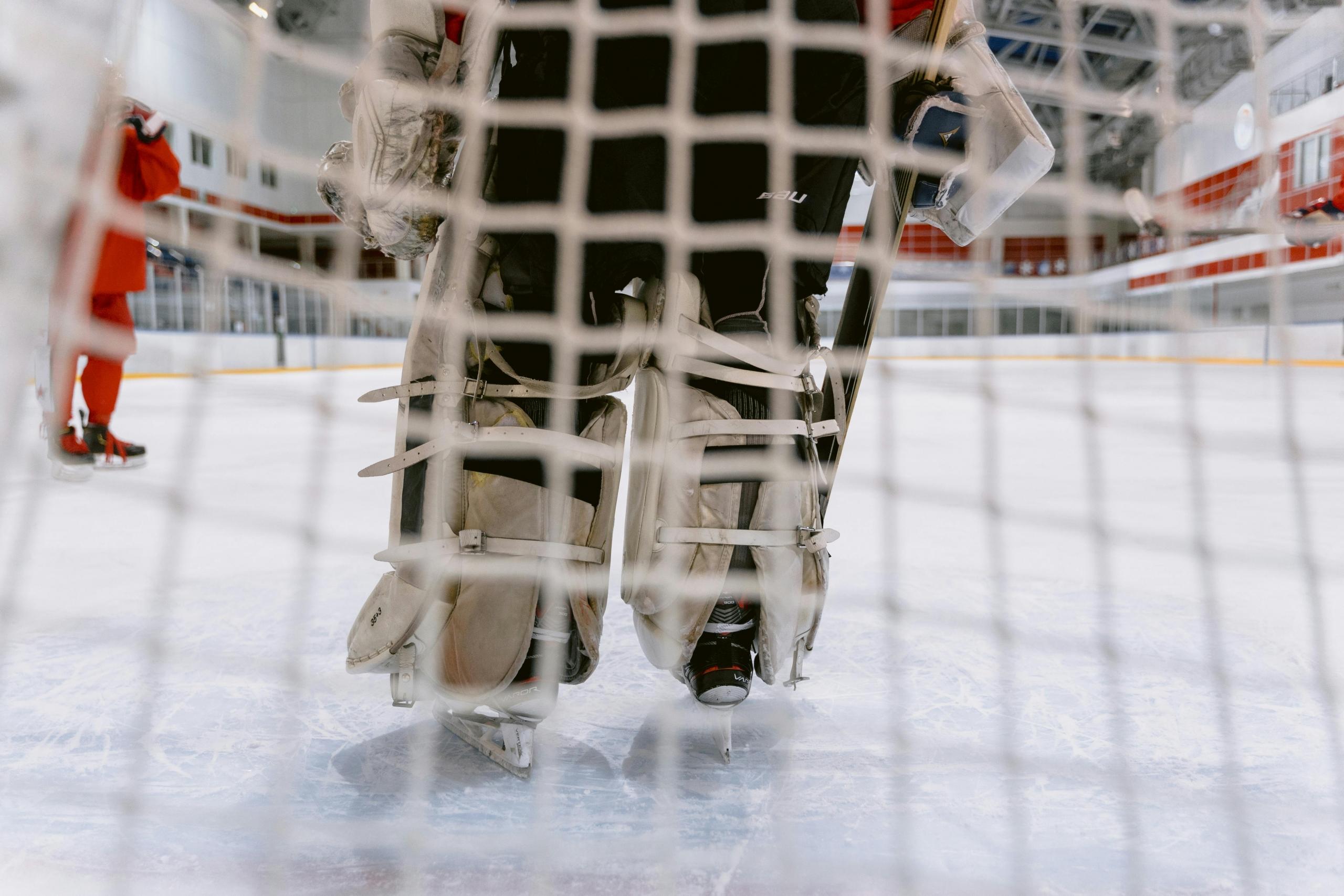 In a view from the bottom of the net, a goalie stands in front of the net in a hockey rink.